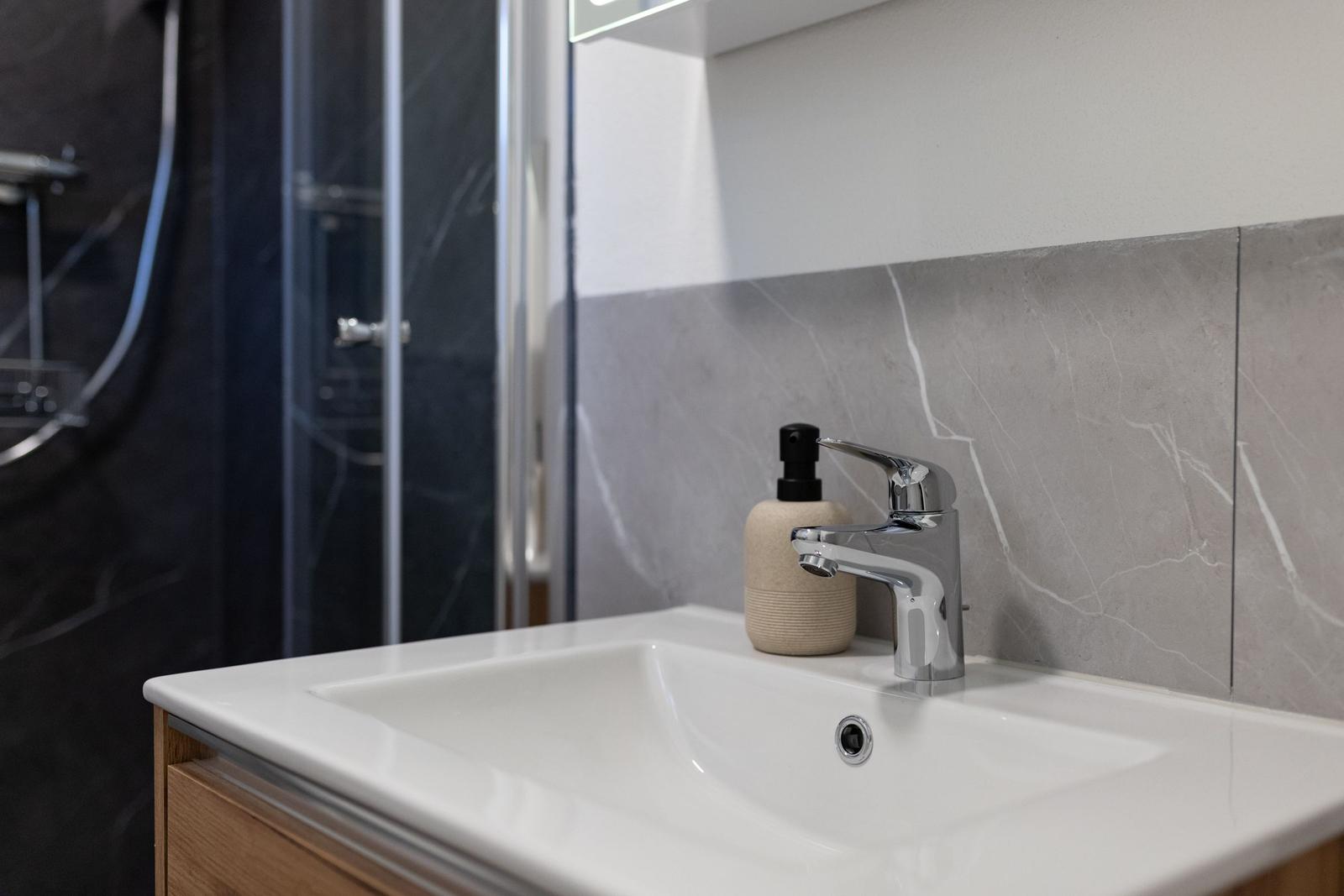 Waschbecken mit Wasserhahn und Seifenspender vor grauem Fliesenspiegel.