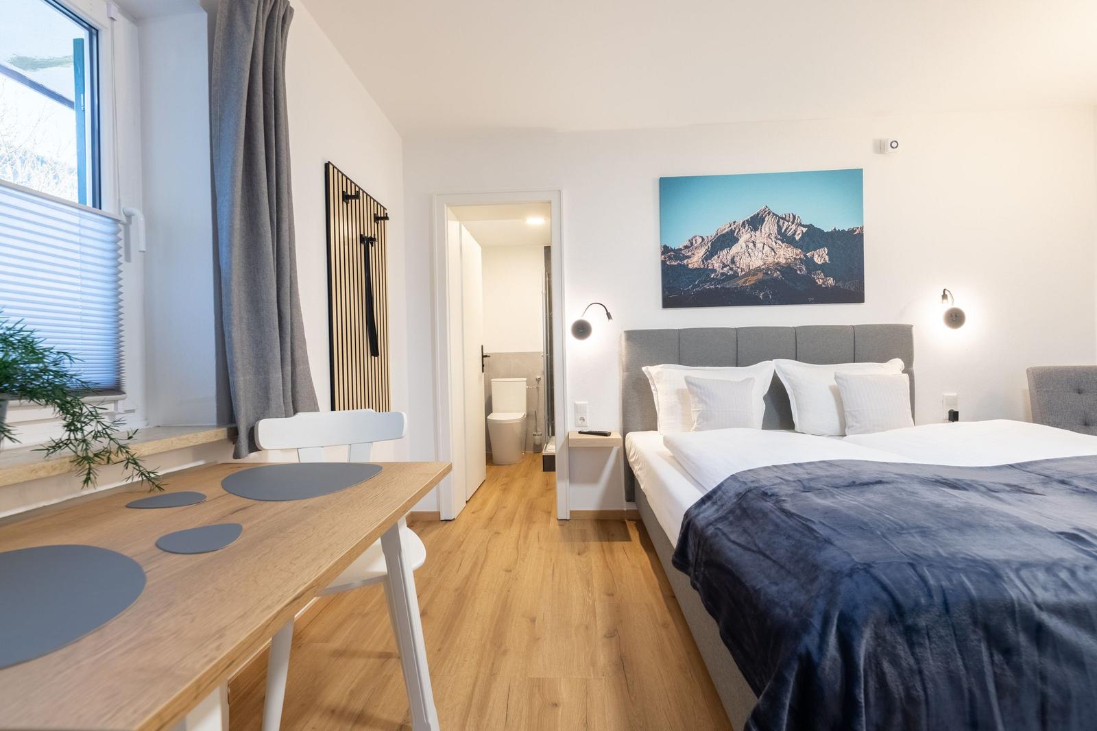 Schlafzimmer mit Doppelbett, Tisch, Fenster und Wandbild mit Bergmotiv.