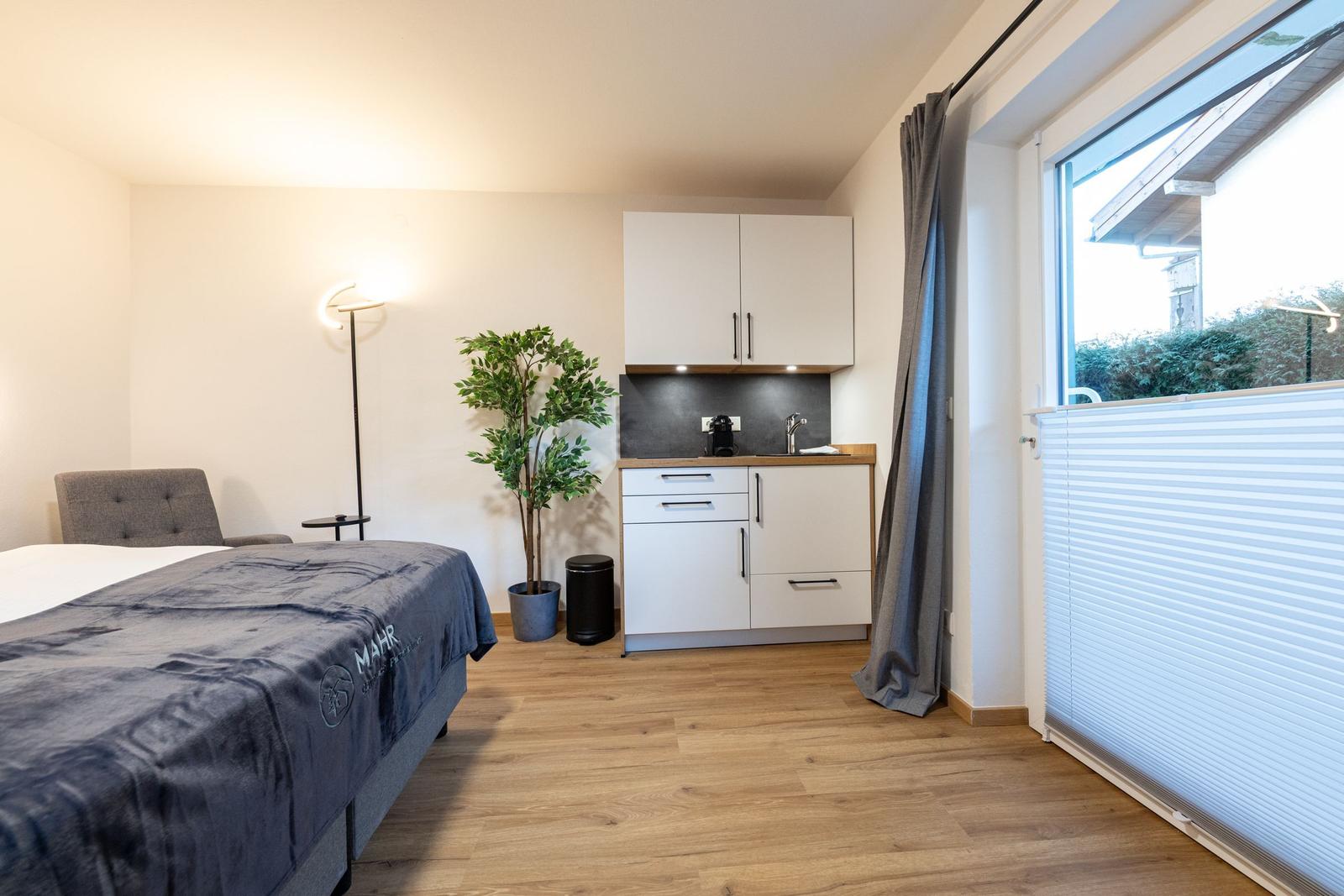 Zimmer mit Bett, kleiner Küchenzeile und Fenster mit Jalousie.