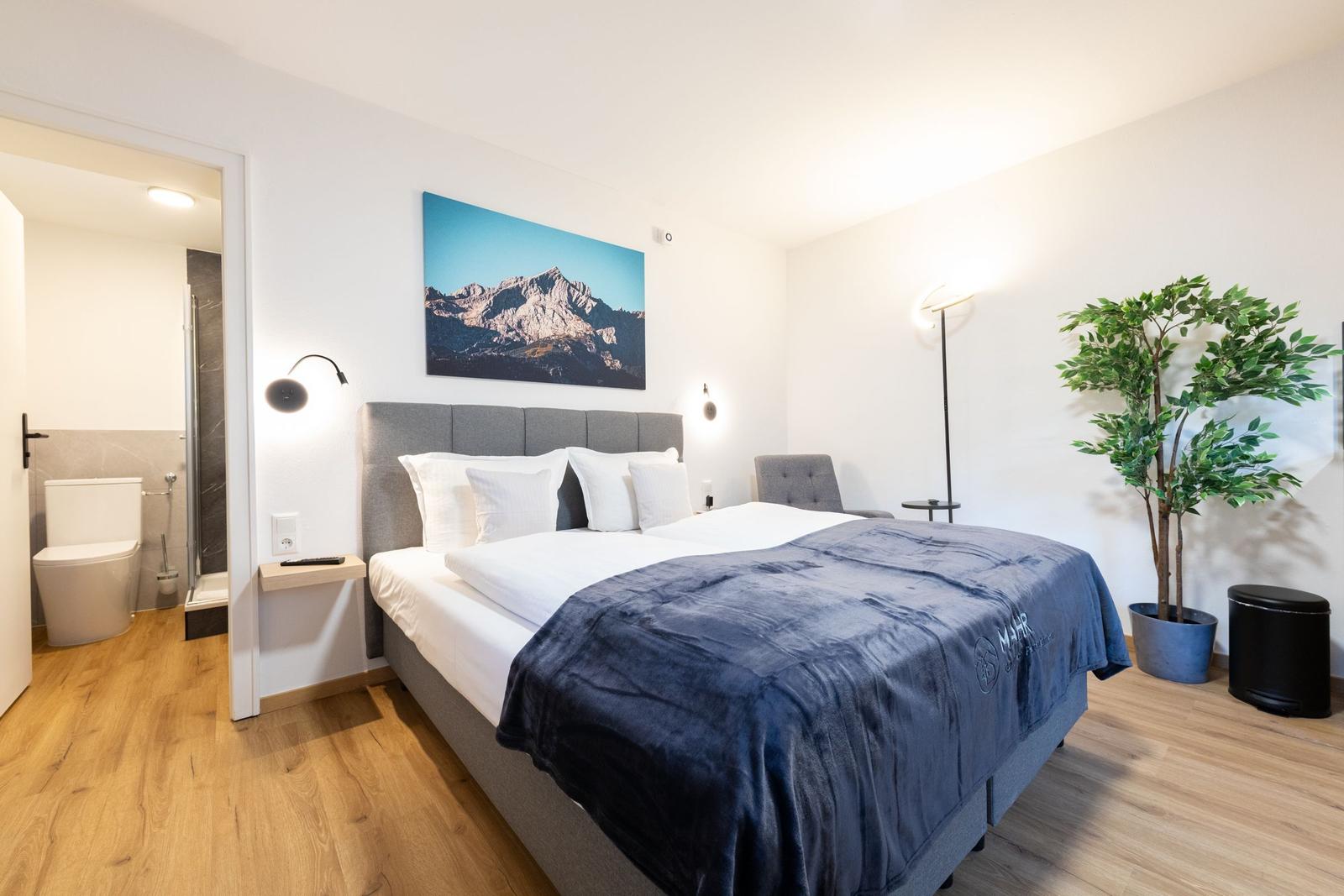 Schlafzimmer mit Doppelbett, Wandbild und Pflanze. Nebenraum mit Toilette und Dusche.