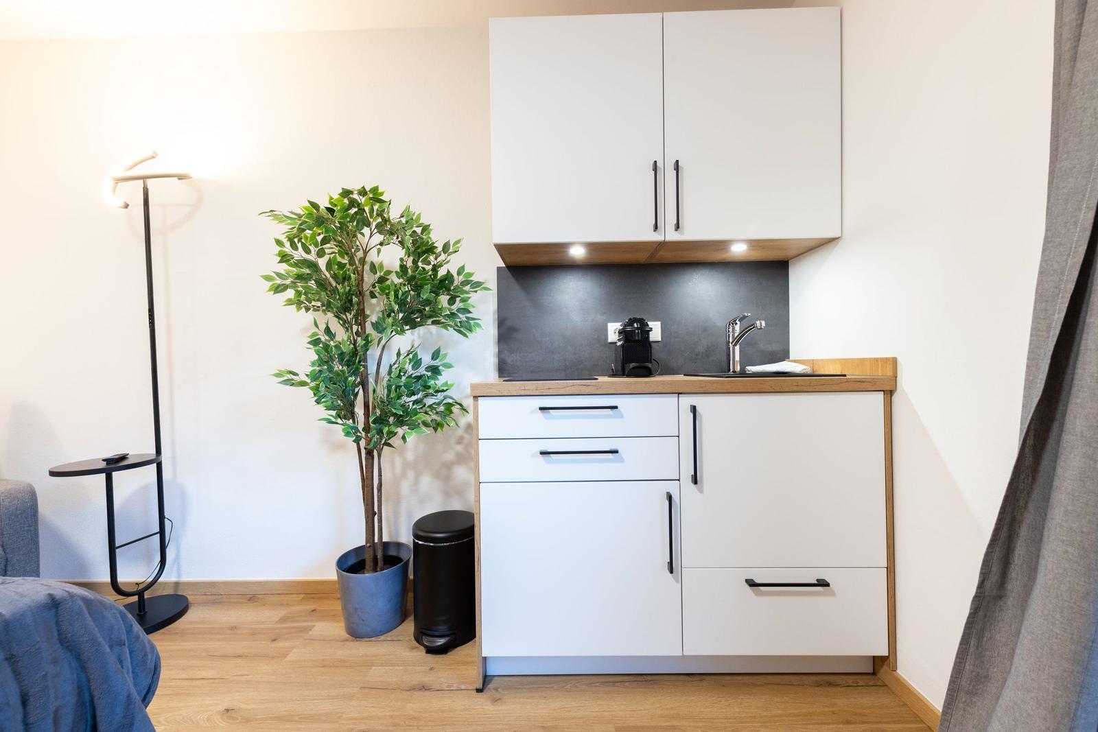 Küche mit weißen Schränken, Holzoberfläche, Spüle und Kaffeemaschine. Pflanze und Stehlampe daneben.