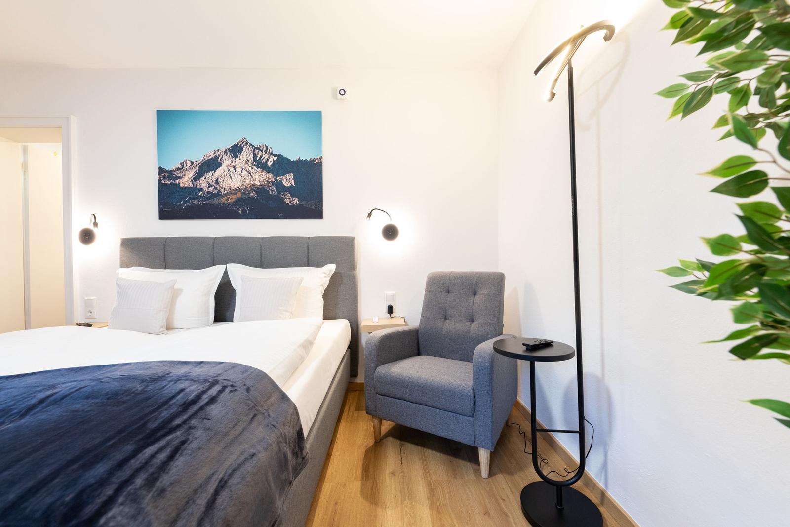 Schlafzimmer mit Bett, Sessel und Wandbild. Holzboden und Pflanze sichtbar.