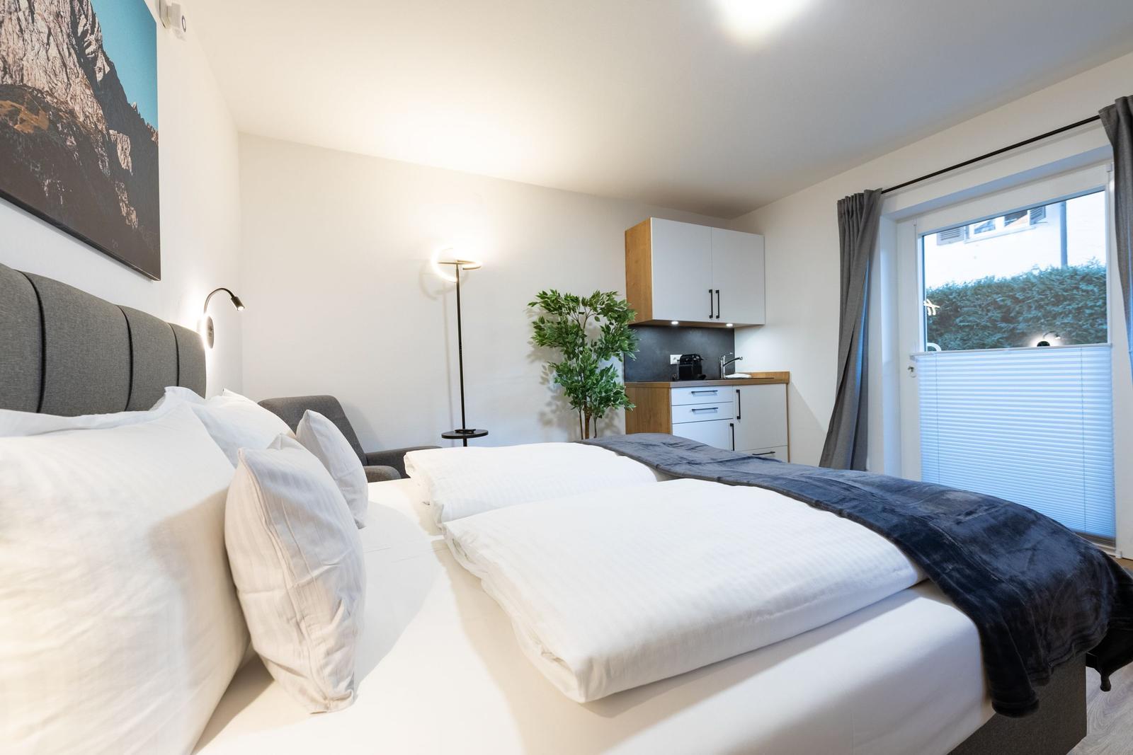 Schlafzimmer mit Doppelbett, Küchenzeile und Fenster mit Jalousie.