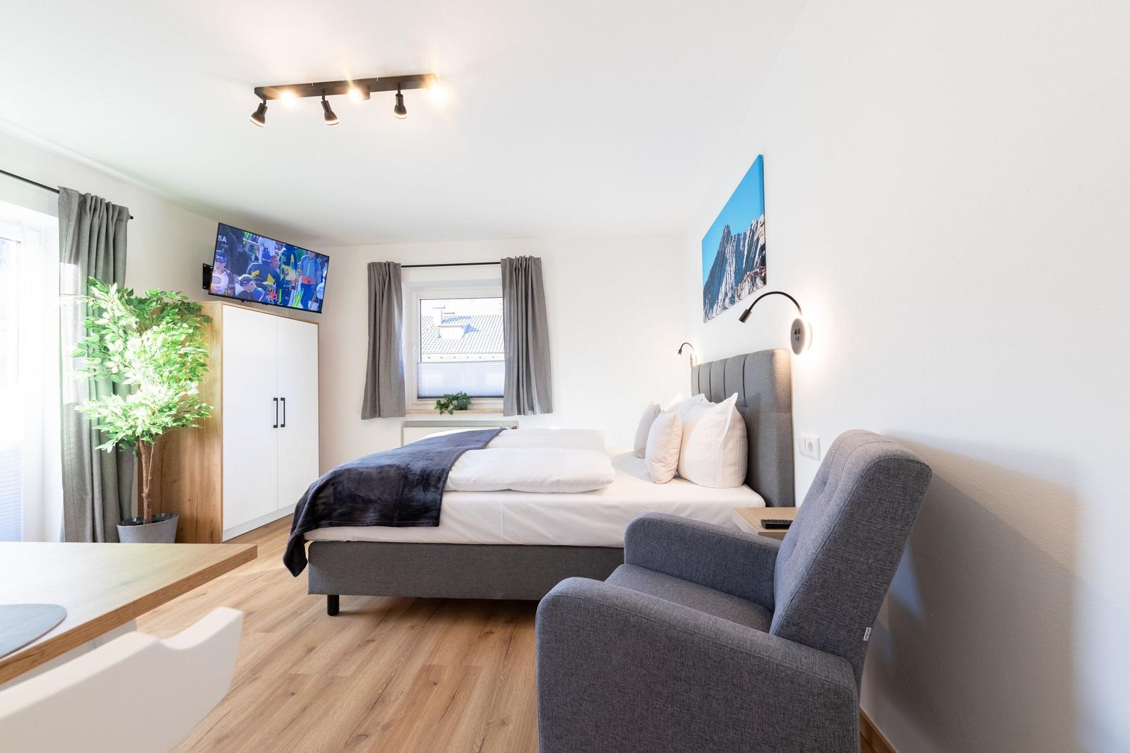 Schlafzimmer mit Bett, Sessel und TV. Fenster mit Vorhängen. Wandbild und Pflanze.