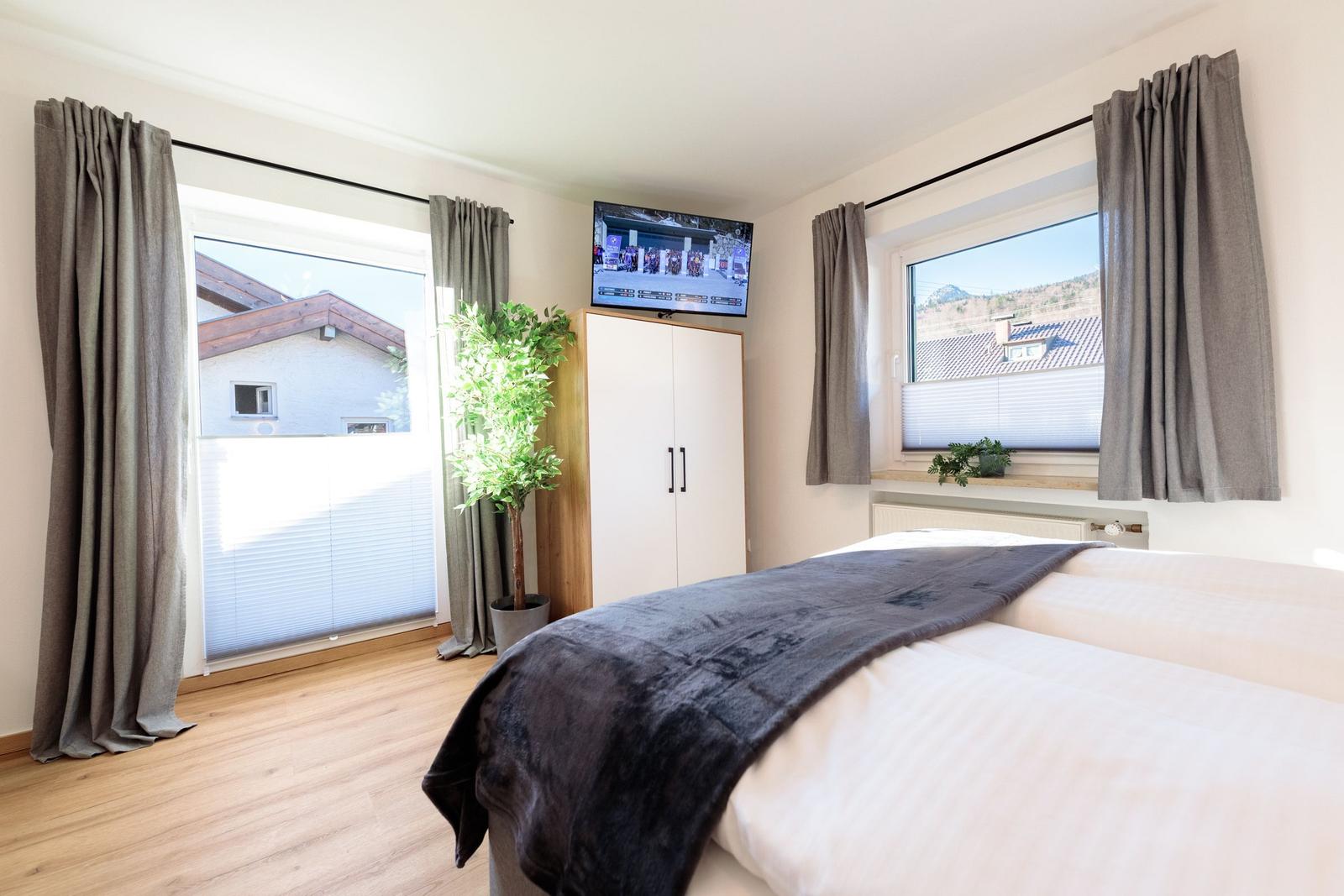 Schlafzimmer mit Bett, TV und zwei Fenstern mit Blick auf die Umgebung.