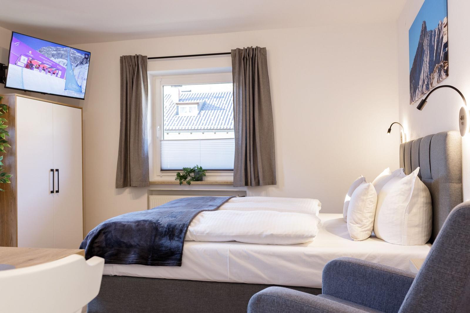 Ein Schlafzimmer mit Bett, TV und Fenster. An der Wand hängt ein Bild.