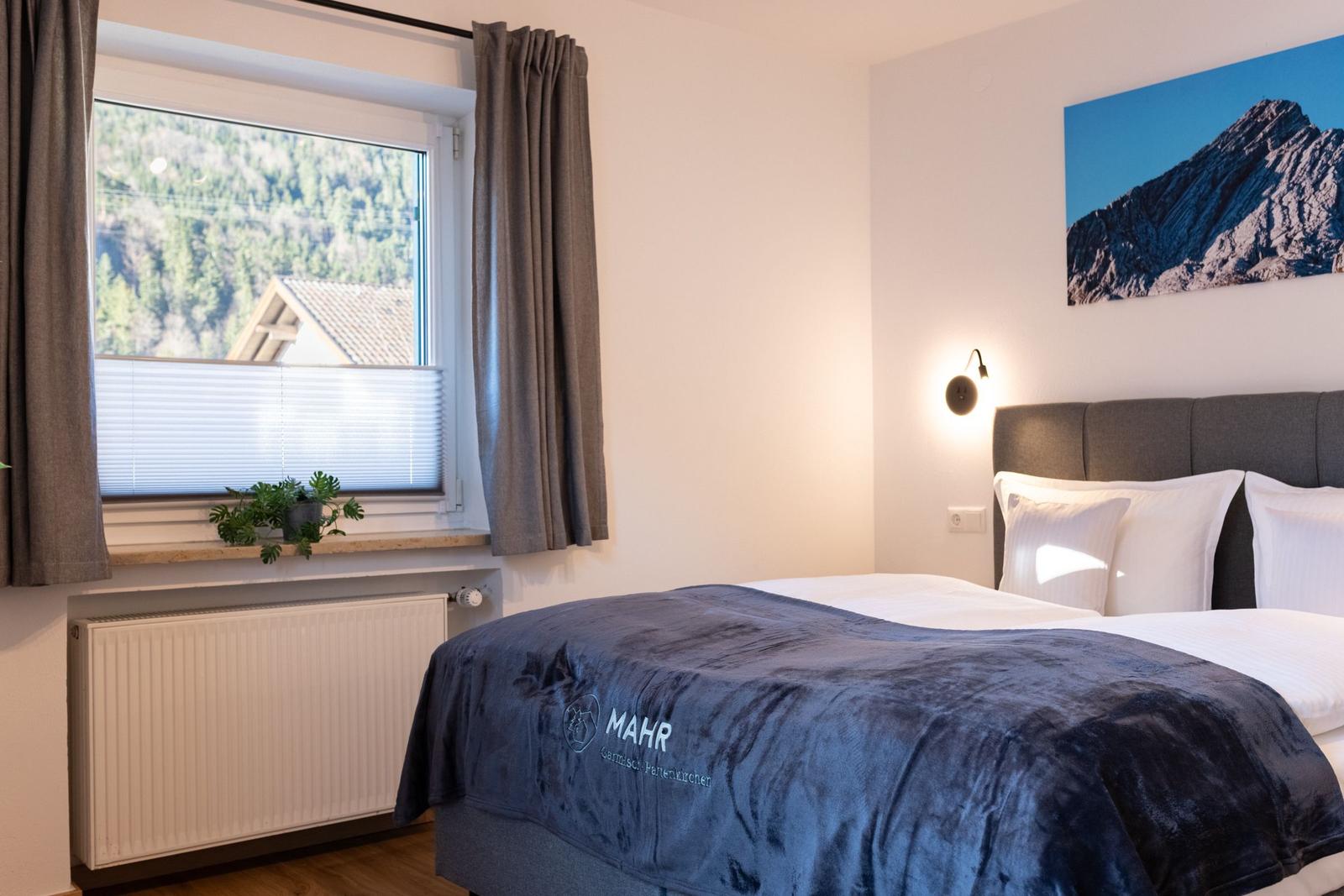 Schlafzimmer mit Bett, Fenster und Wandbild. Blick auf Wald und Haus.