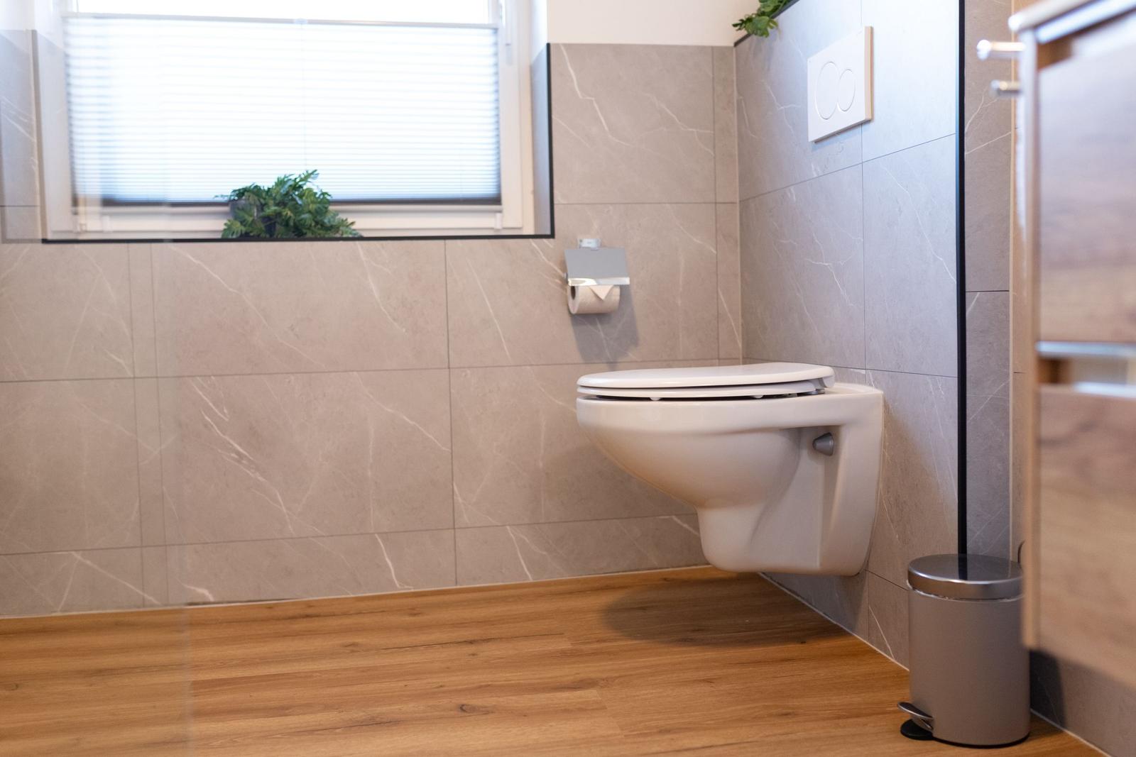 WC mit Wandmontage, Holzboden und Fenster mit Jalousie.