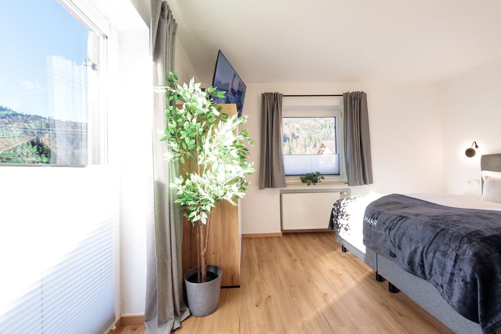 Schlafzimmer mit Bett, Fenster und Zimmerpflanze. Holzboden und Blick auf die Berge.