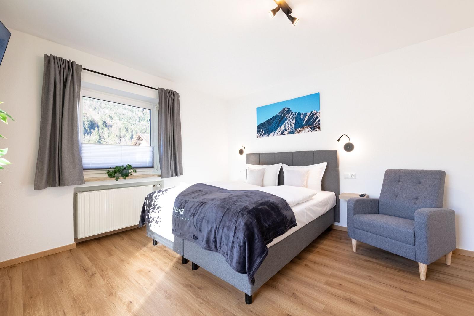 Ein Schlafzimmer mit Bett, Sessel und Fensterblick auf Berge.