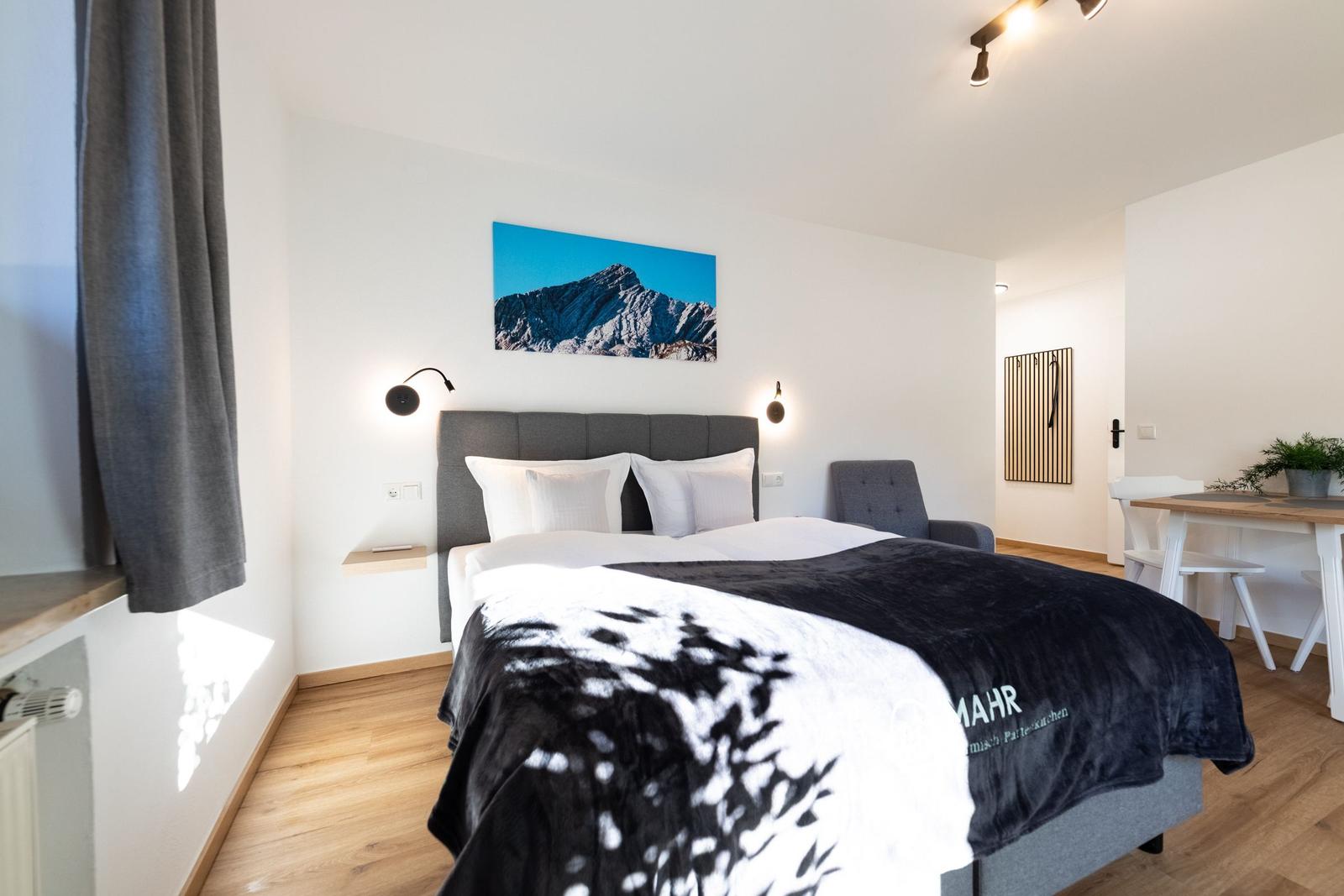 Schlafzimmer mit Bett, Wandbild und Sitzgelegenheit. Holzboden und Fenster mit Vorhang.