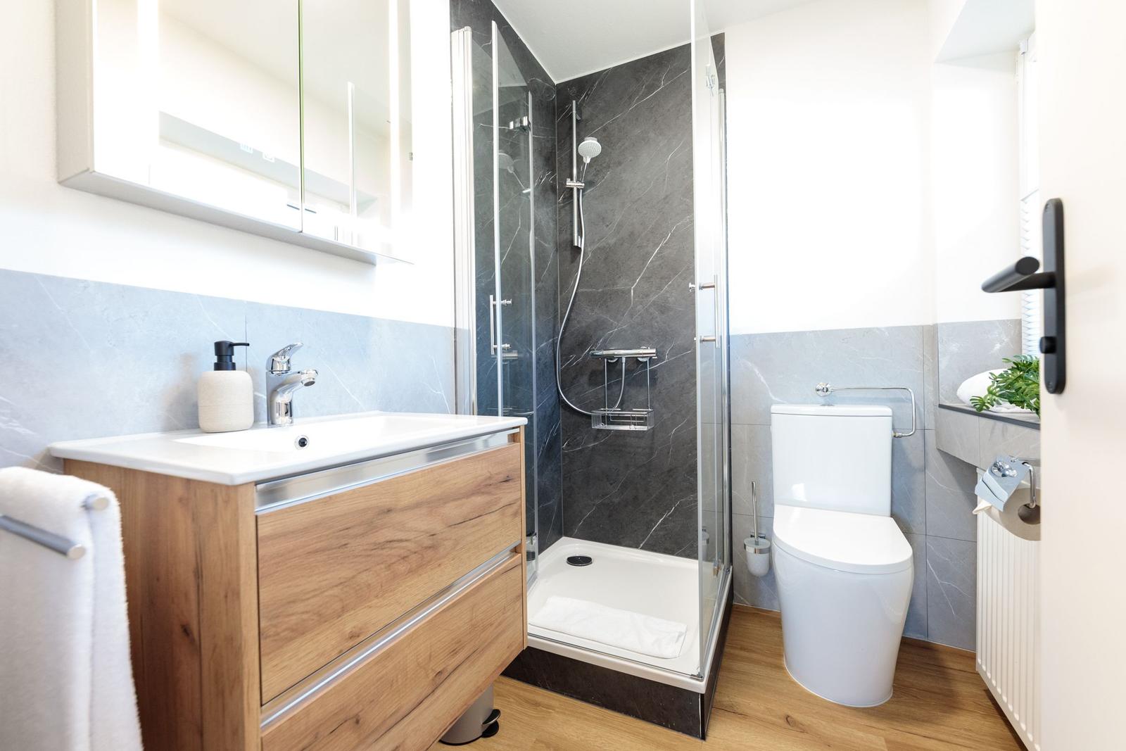 Badezimmer mit Dusche, Waschbecken und Toilette. Holzoptik und graue Fliesen.