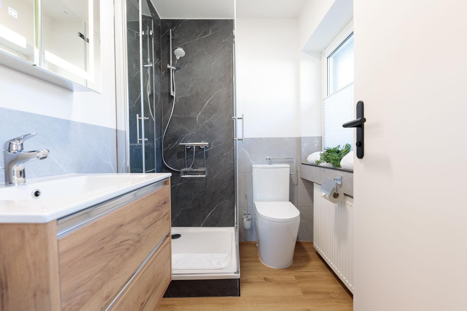 Badezimmer mit Dusche, Waschbecken und Toilette. Holzboden und graue Fliesen.