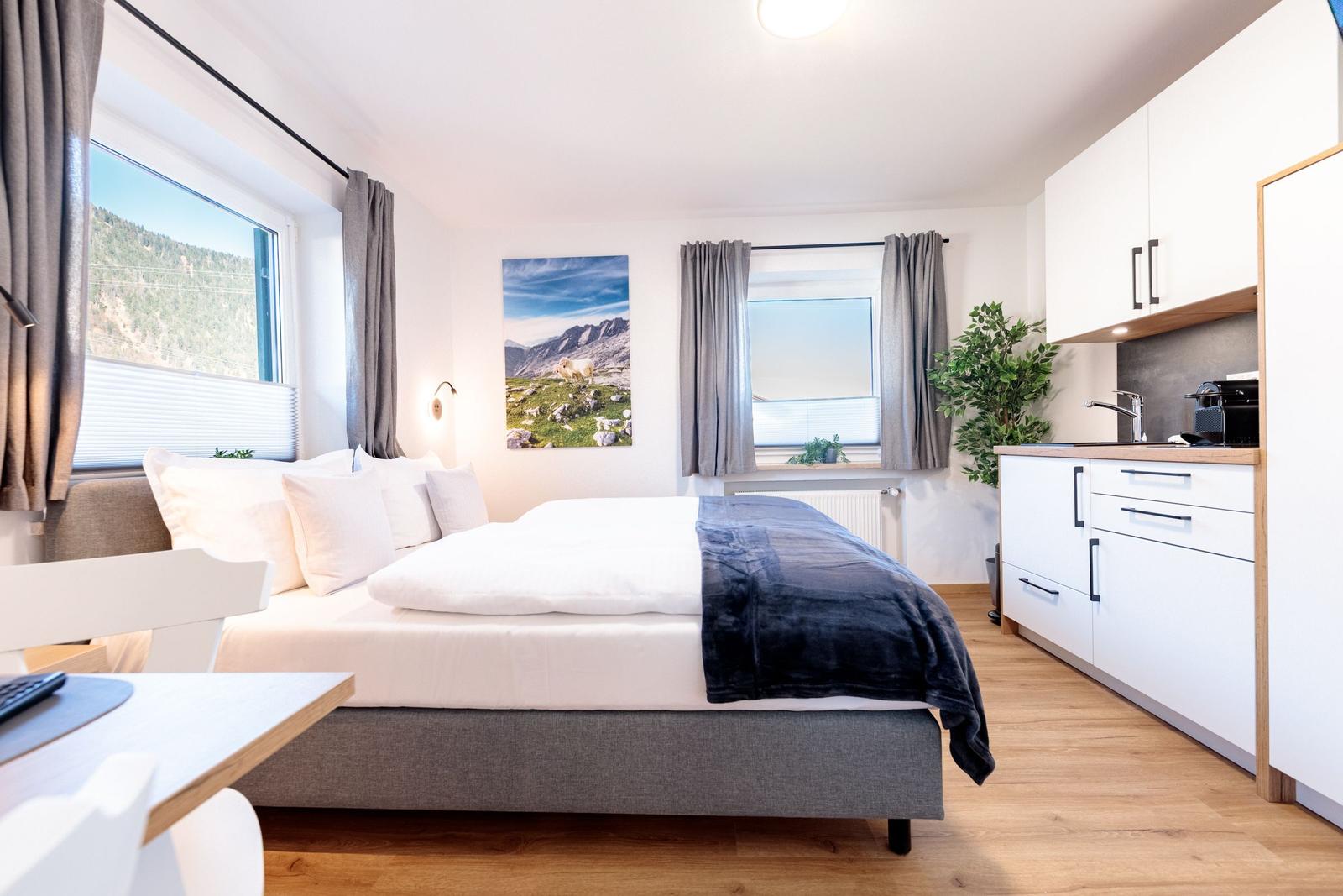 Schlafzimmer mit Bett, Fenster und Küchenbereich. Wandbild zeigt Berglandschaft.