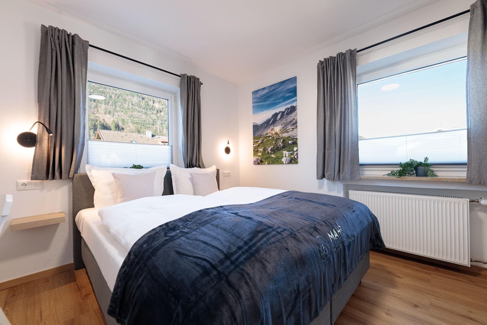 Schlafzimmer mit Doppelbett, zwei Fenstern und Wandbild. Holzboden und graue Vorhänge.