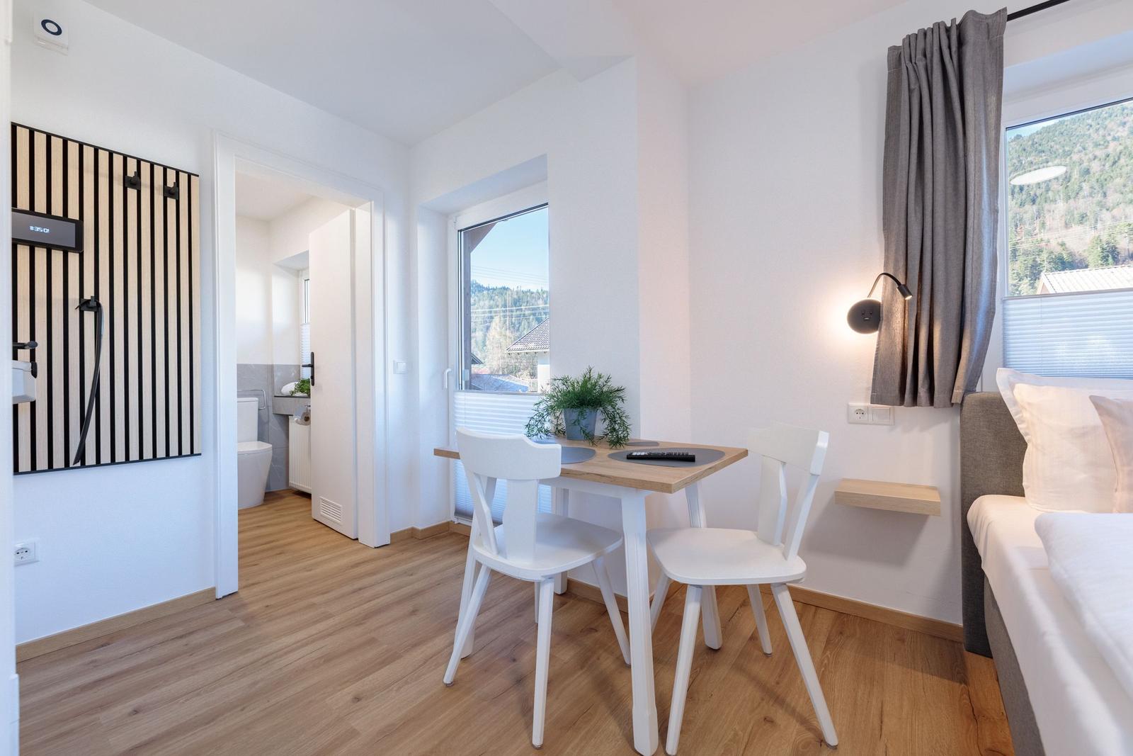 Zimmer mit Tisch, Stühlen, Bett und Bad. Fenster mit Blick auf Wald.