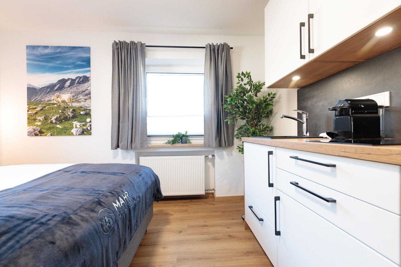 Schlafzimmer mit Bett, Fenster und Küchenzeile mit Arbeitsplatte und Kaffeemaschine.