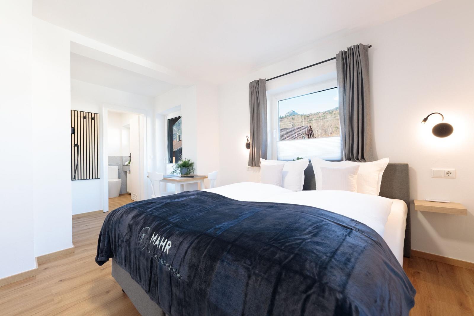 Schlafzimmer mit Bett, Fenster und Blick auf Berge. Nebenraum mit Toilette und Dusche.