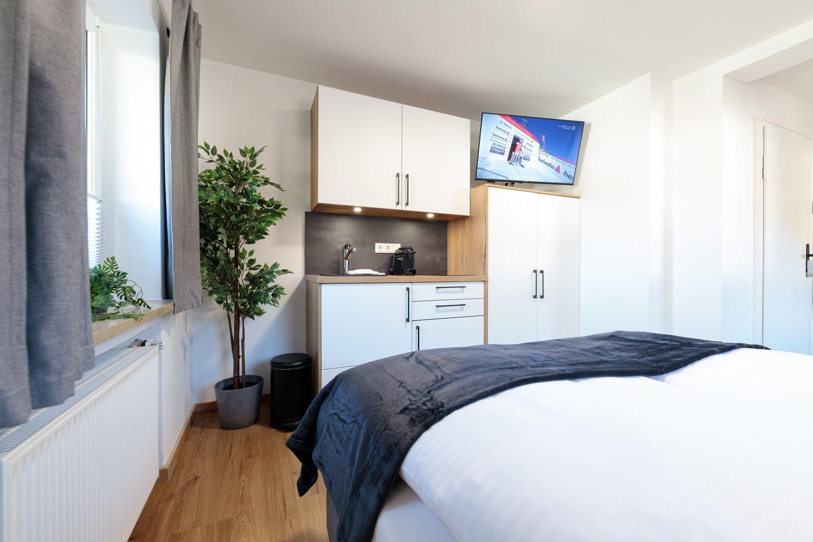 Schlafzimmer mit Bett, Kleiderschrank und Küchenzeile. Fenster mit Vorhängen und Zimmerpflanze.