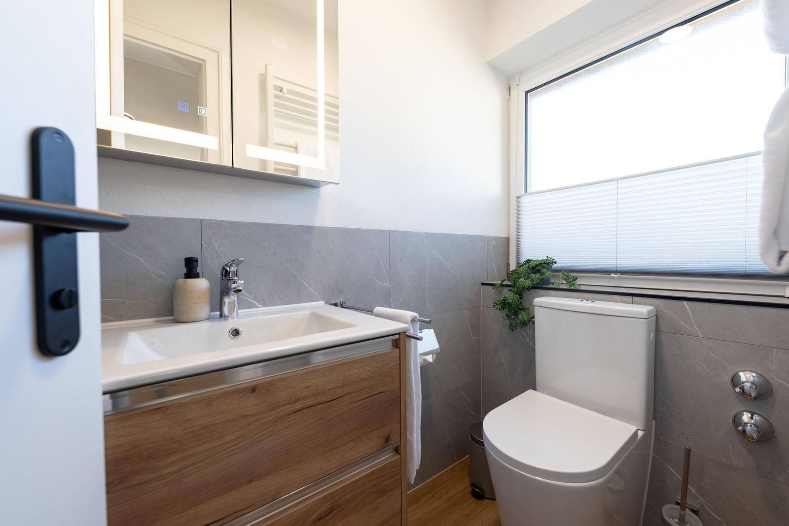 Badezimmer mit Waschbecken, Spiegel und Toilette. Fenster mit Jalousie und Pflanze.