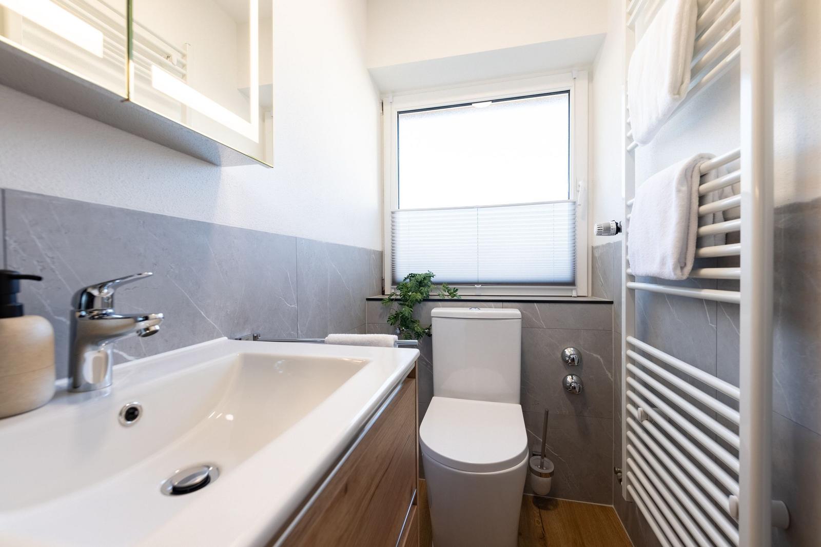 Badezimmer mit Waschbecken, Toilette und Heizung. Fenster mit Jalousie und Pflanze.