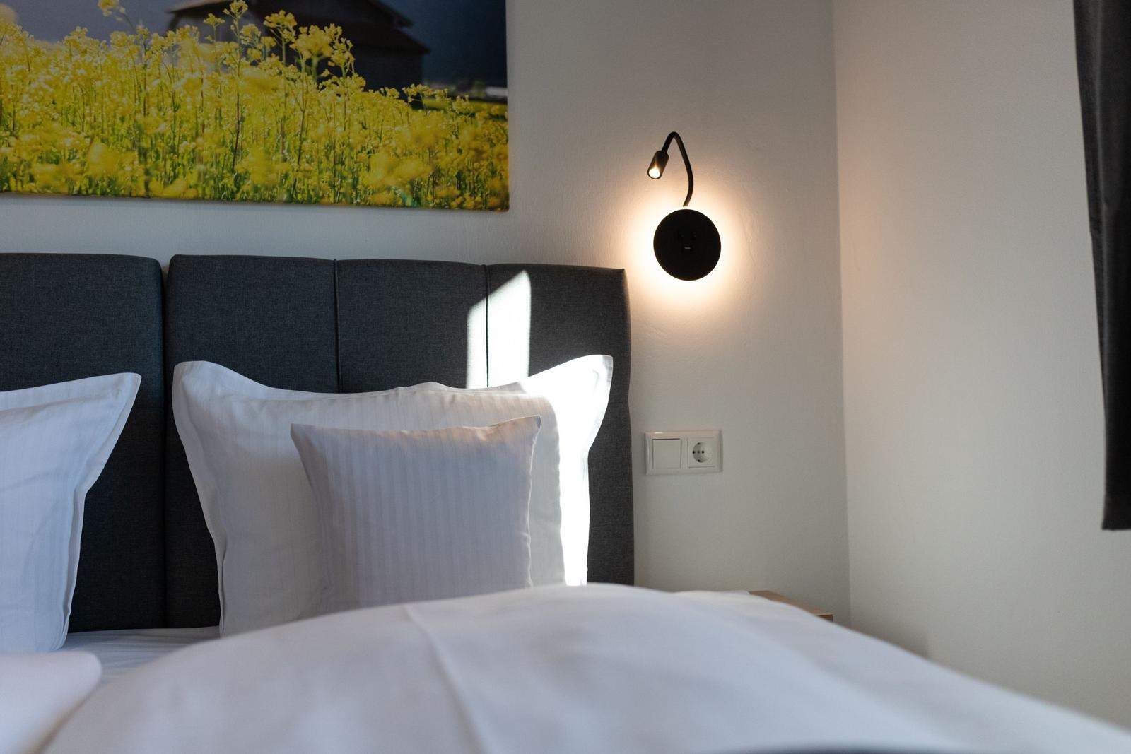 Schlafzimmer mit Bett, Kopfkissen und Wandlampe. Wandbild zeigt gelbe Blumen.