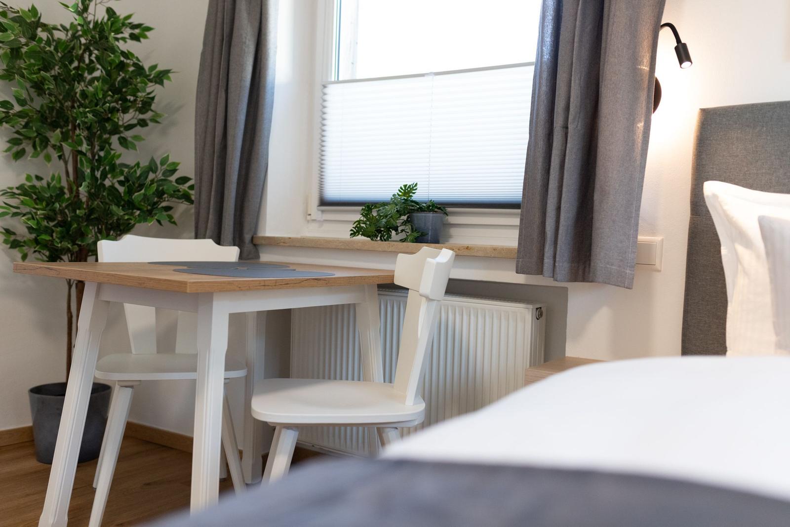 Ein Bett mit grauem Kopfteil, ein weißer Tisch mit zwei Stühlen und eine Fensterbank mit Pflanzen.