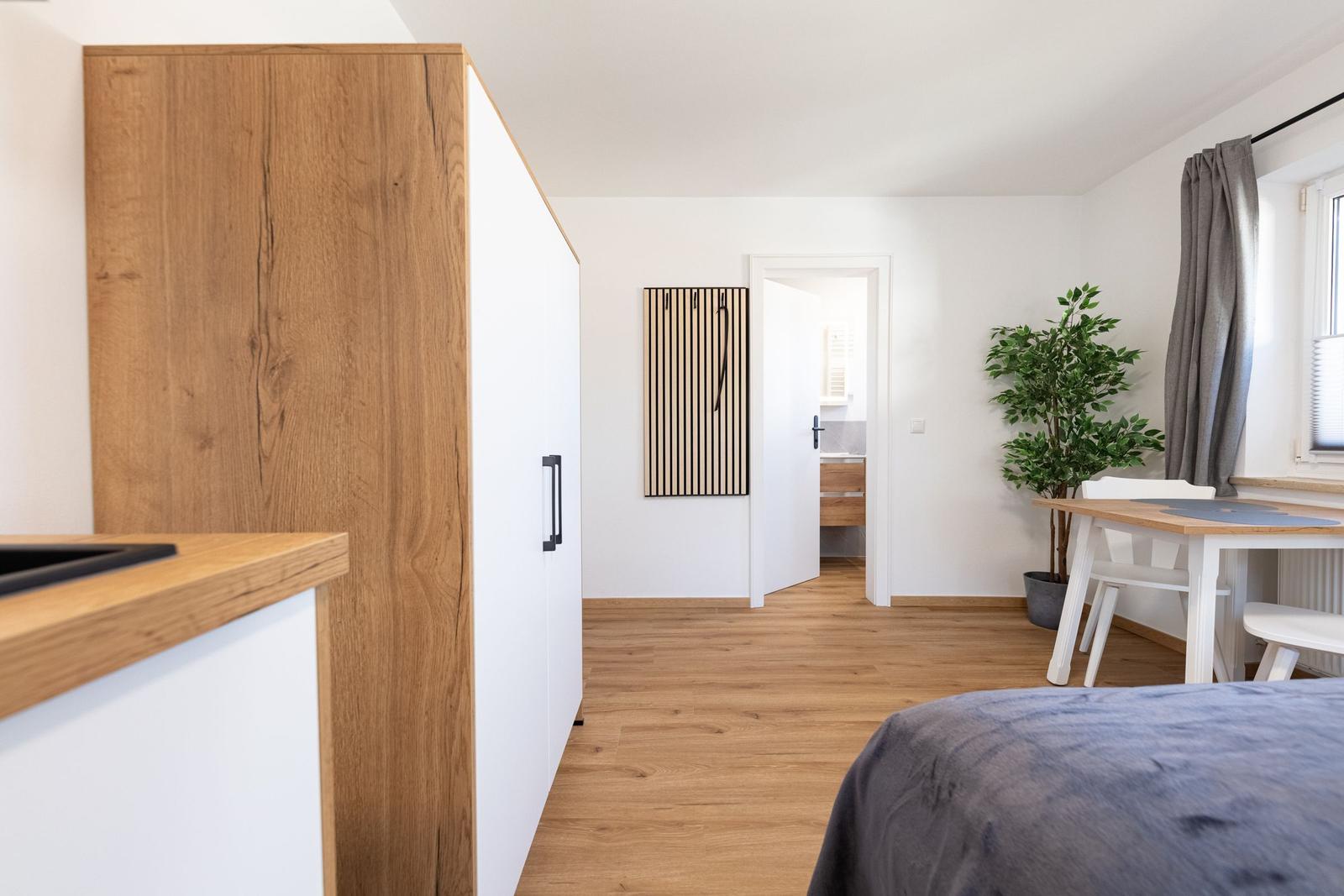 Ein Raum mit Holzboden, Bett, Tisch, Stühlen und Pflanze. Tür führt ins Bad.