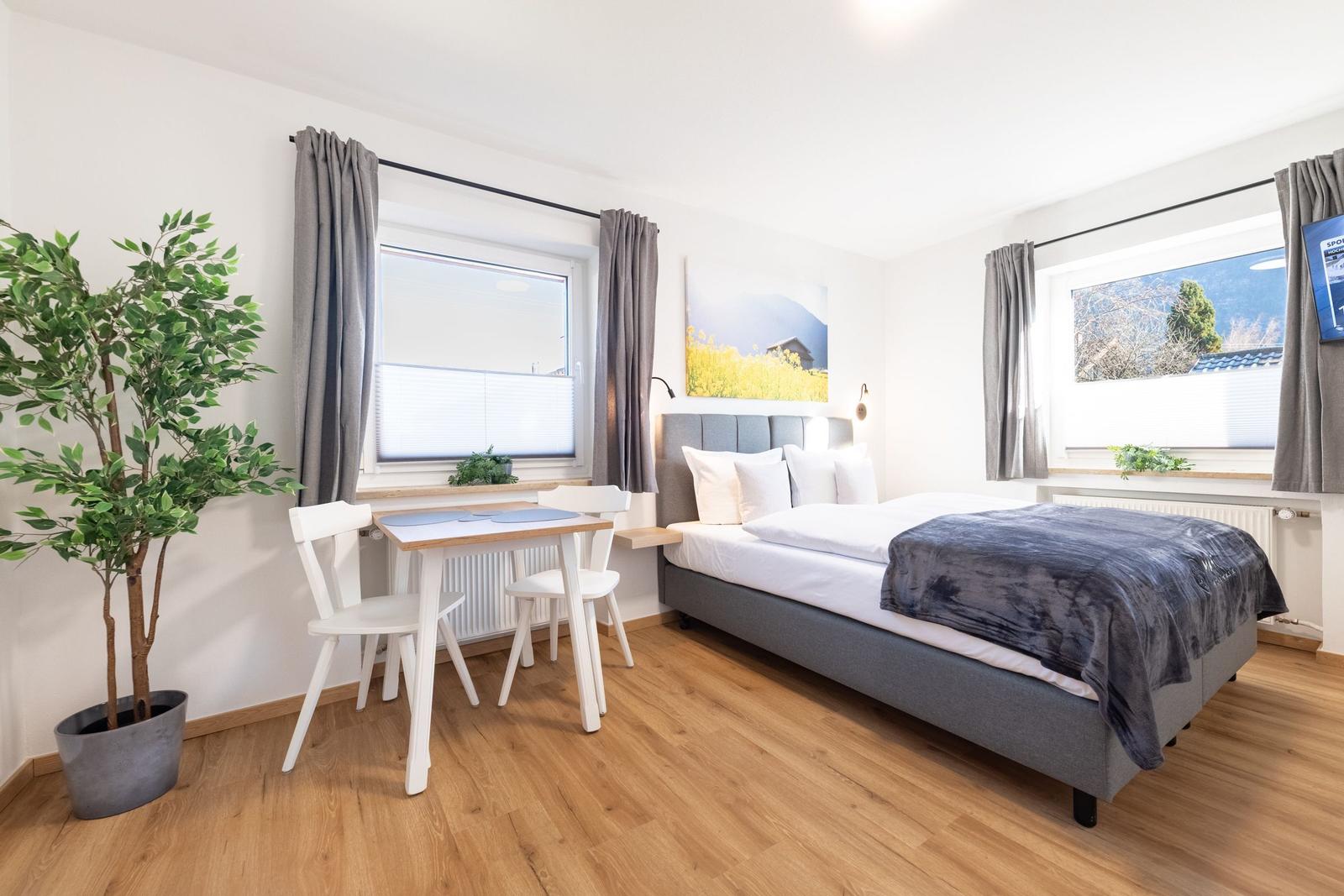 Ein Schlafzimmer mit Bett, Tisch und Stühlen, Fenstern und Zimmerpflanze.