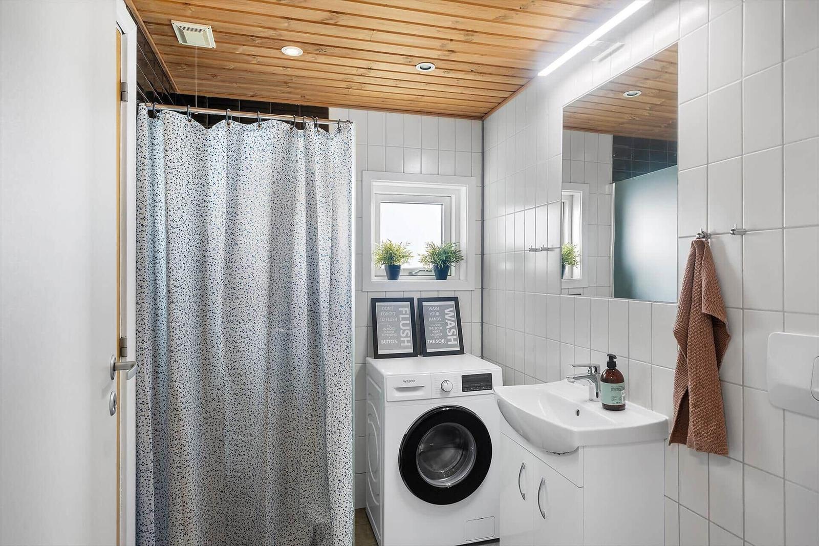Badezimmer mit Dusche, Waschmaschine und Waschbecken. Holzdecke und weiße Fliesen.