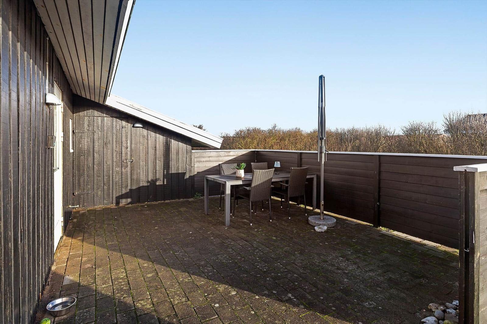 Terrasse mit Tisch, Stühlen und Sonnenschutz. Holzwand und Pflastersteinboden.
