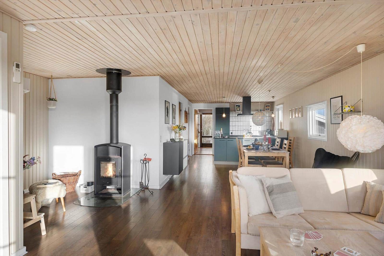Wohnzimmer mit Kamin, Sofa und Essbereich. Holzdecke und -boden. Küche im Hintergrund.
