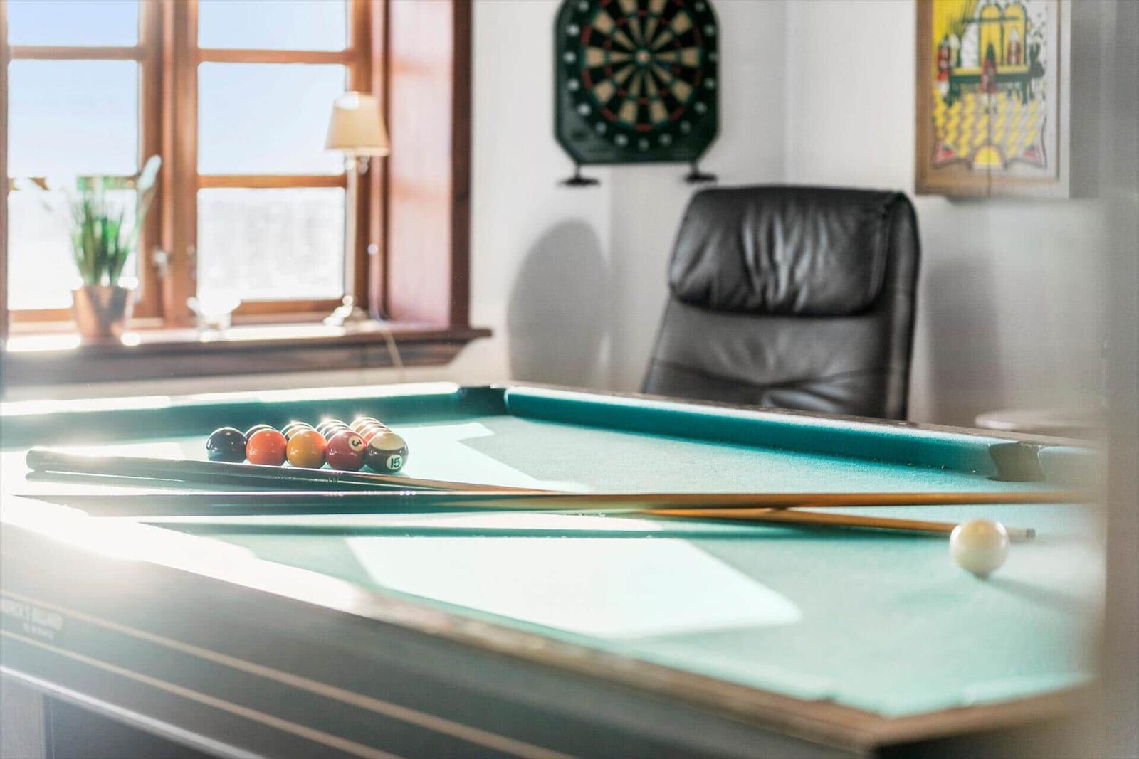 Billiard table with balls and cues. Dartboard and leather chair in background.
