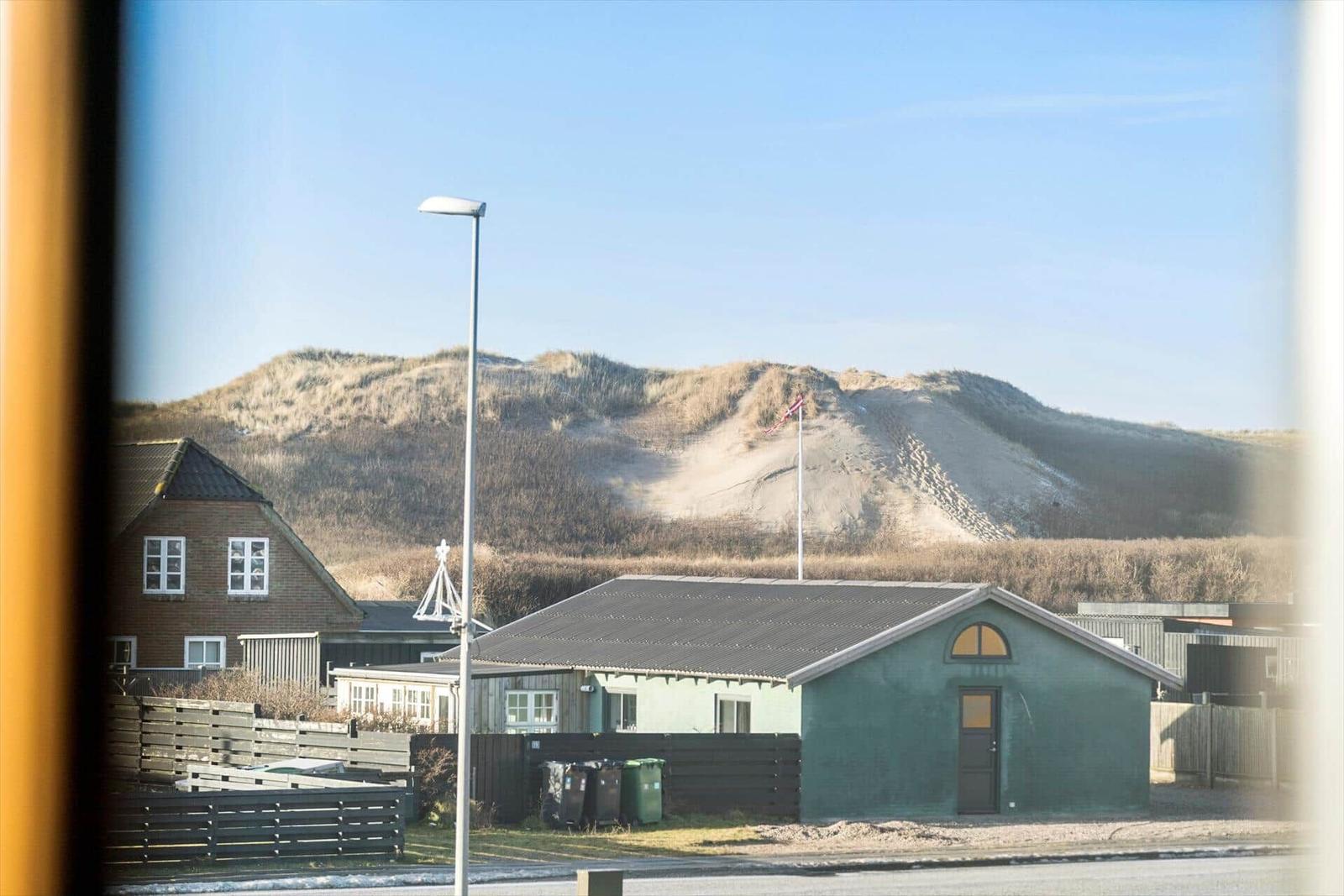 From a window, you see a green cabin, a road, and dunes with a flag.