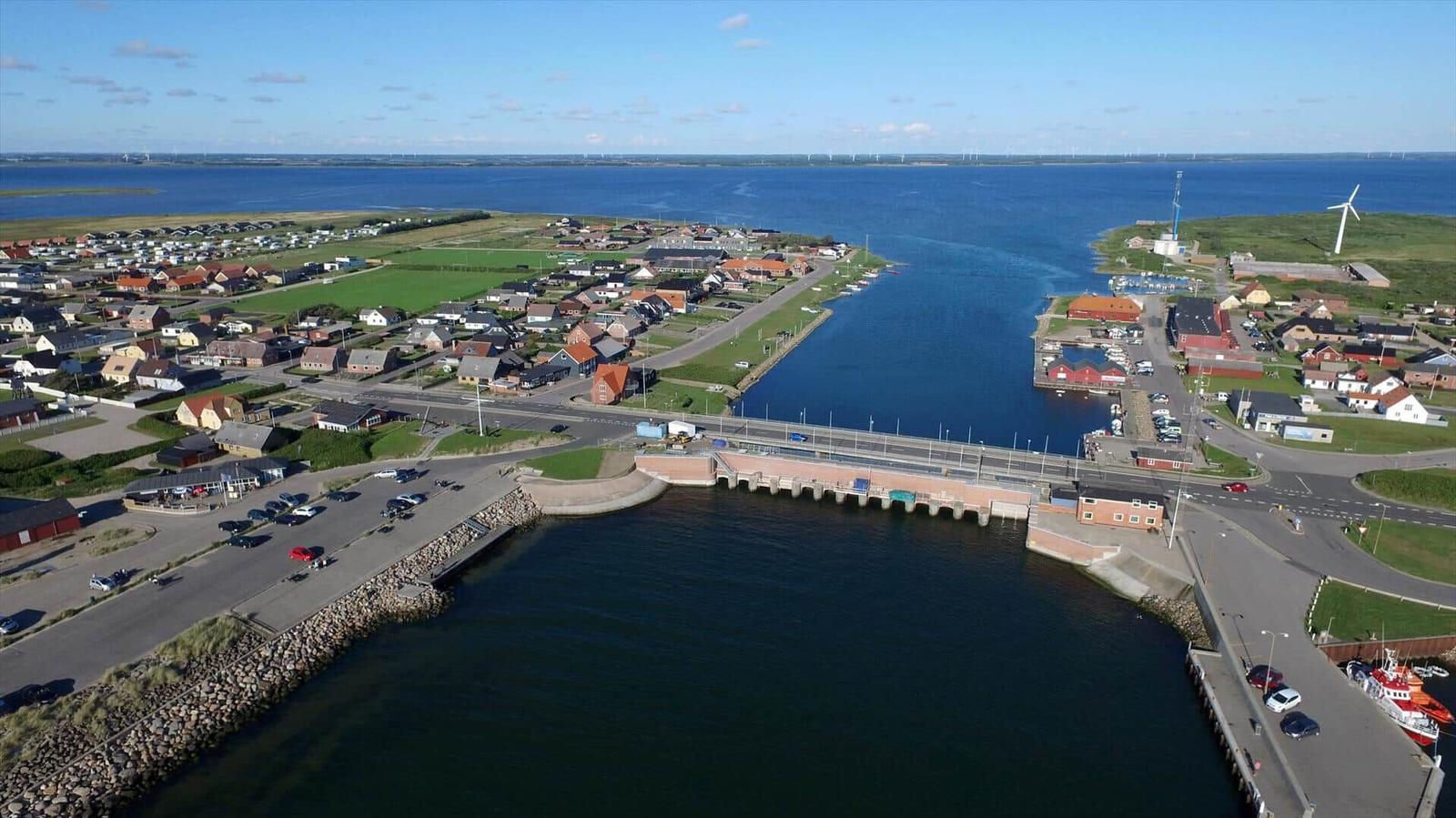 Aerial view of a coastal town with harbor, bridge, and wind turbines in the distance.
