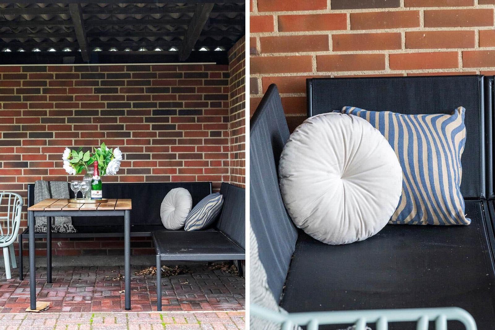 Terrassenbereich mit Sitzbank, Tisch und Kissen vor Backsteinwand.