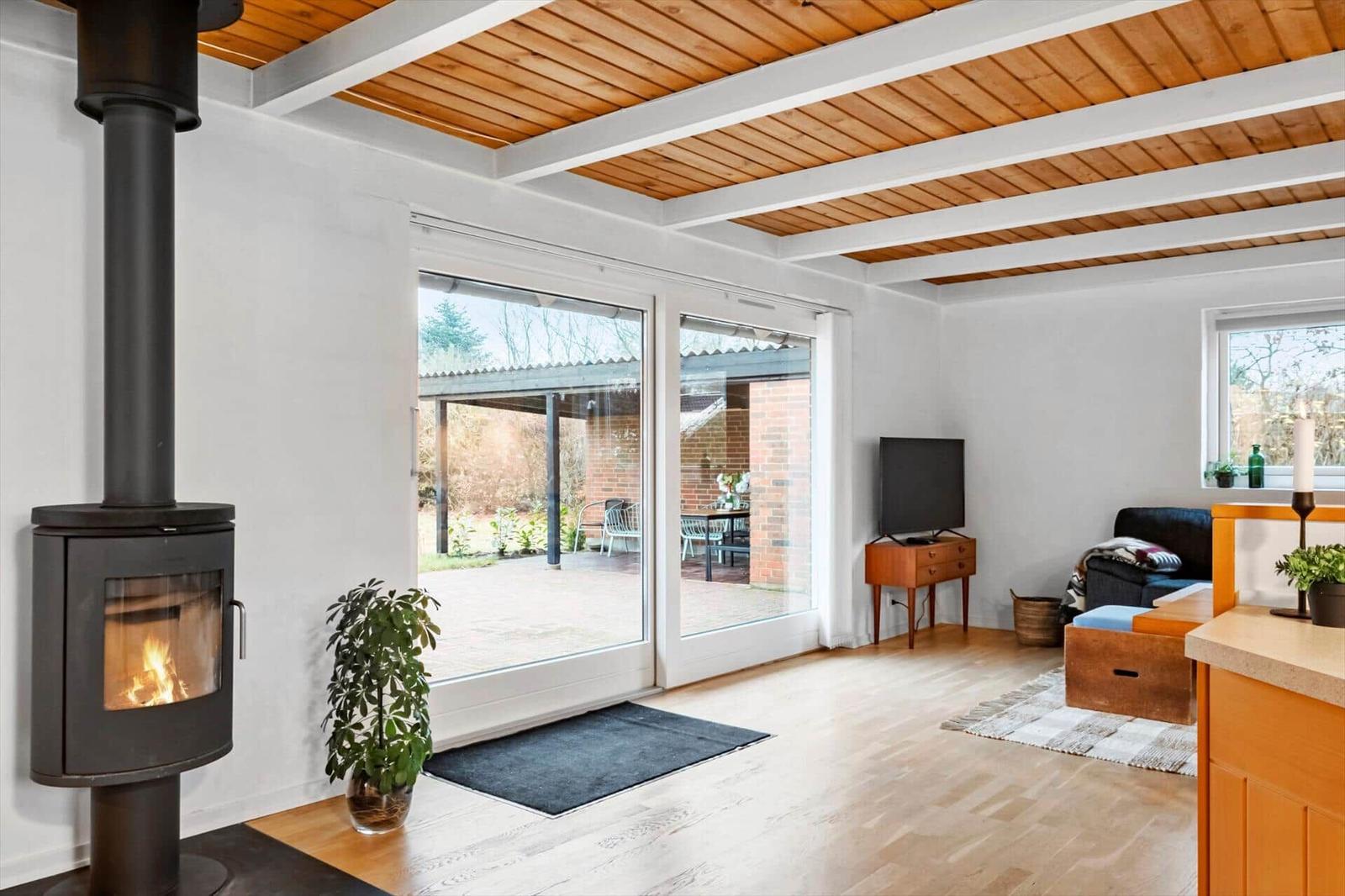 Wohnbereich mit Kamin, Glasfront und Holzboden. Fernseher, Sofa und Küchenzeile sichtbar.