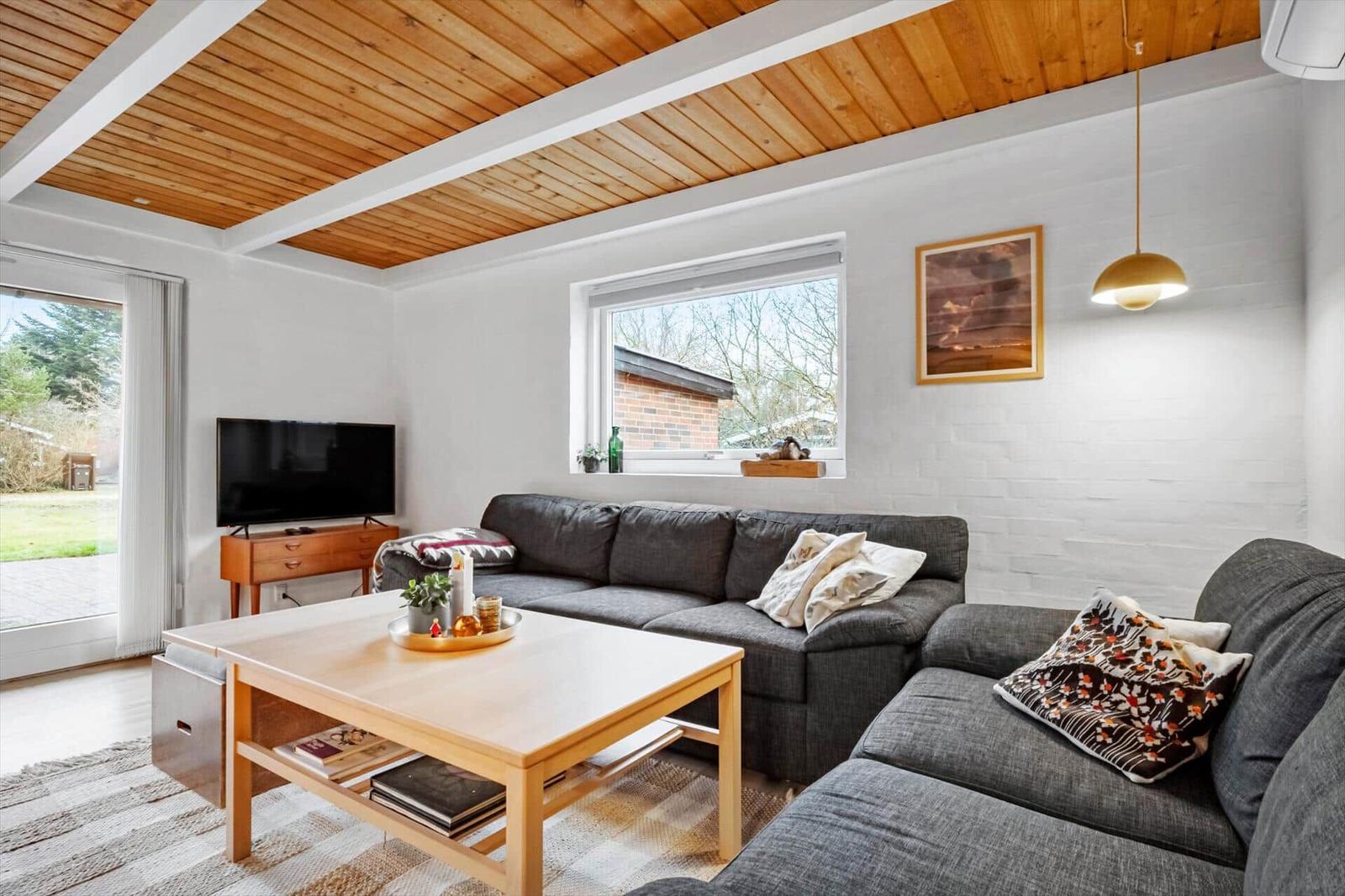 Wohnzimmer mit grauem Sofa, Holztisch und Fernseher. Holzdecke und Fenster mit Blick nach draußen.