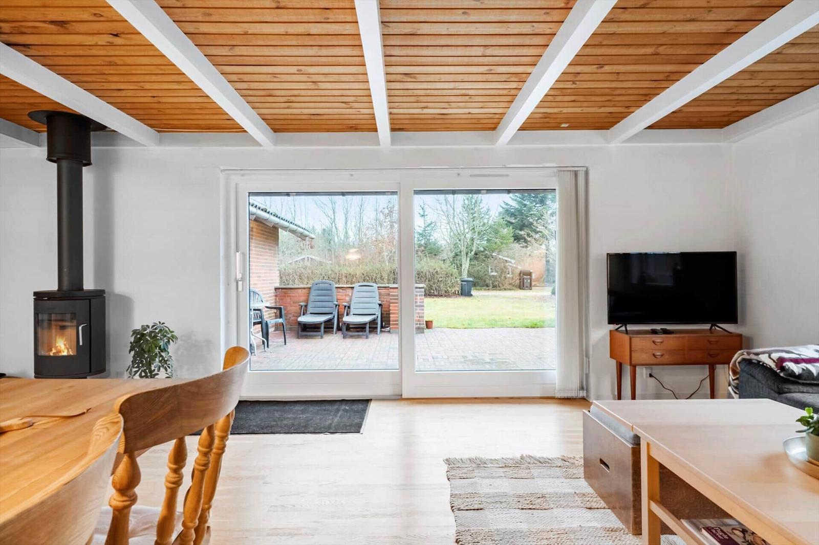 Wohnraum mit Holzdecke, Kamin, TV und Blick auf Terrasse mit Garten.