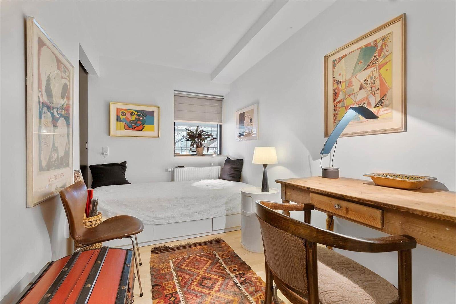 Bedroom with bed, desk, and wall art. Wooden chairs and rug on light walls.