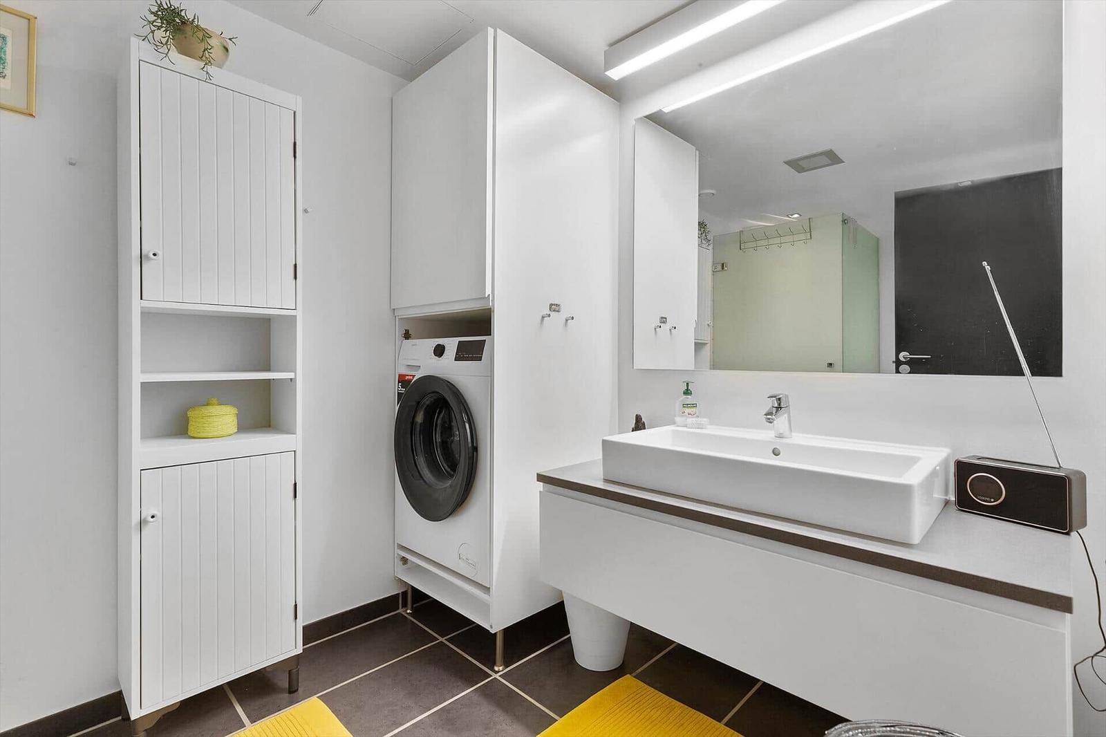 Bathroom with washing machine, mirror, and sink. Yellow rugs and white cabinets.