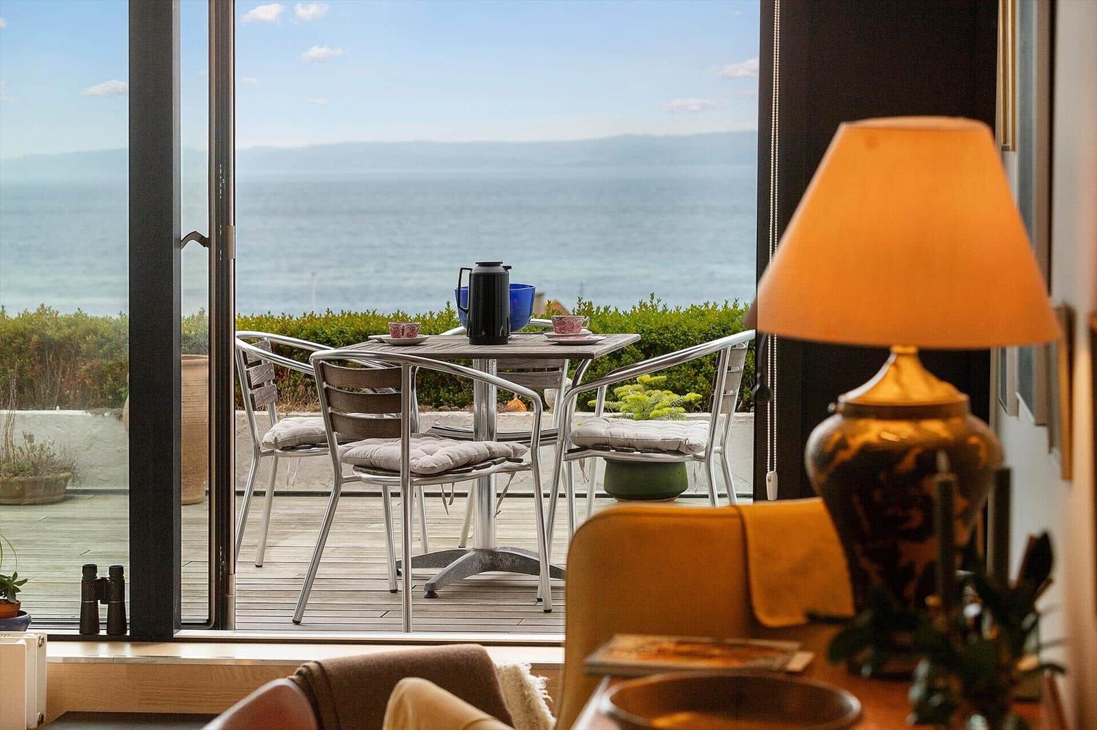 Terrace with table and chairs offers sea view. In foreground lamp and seating furniture.