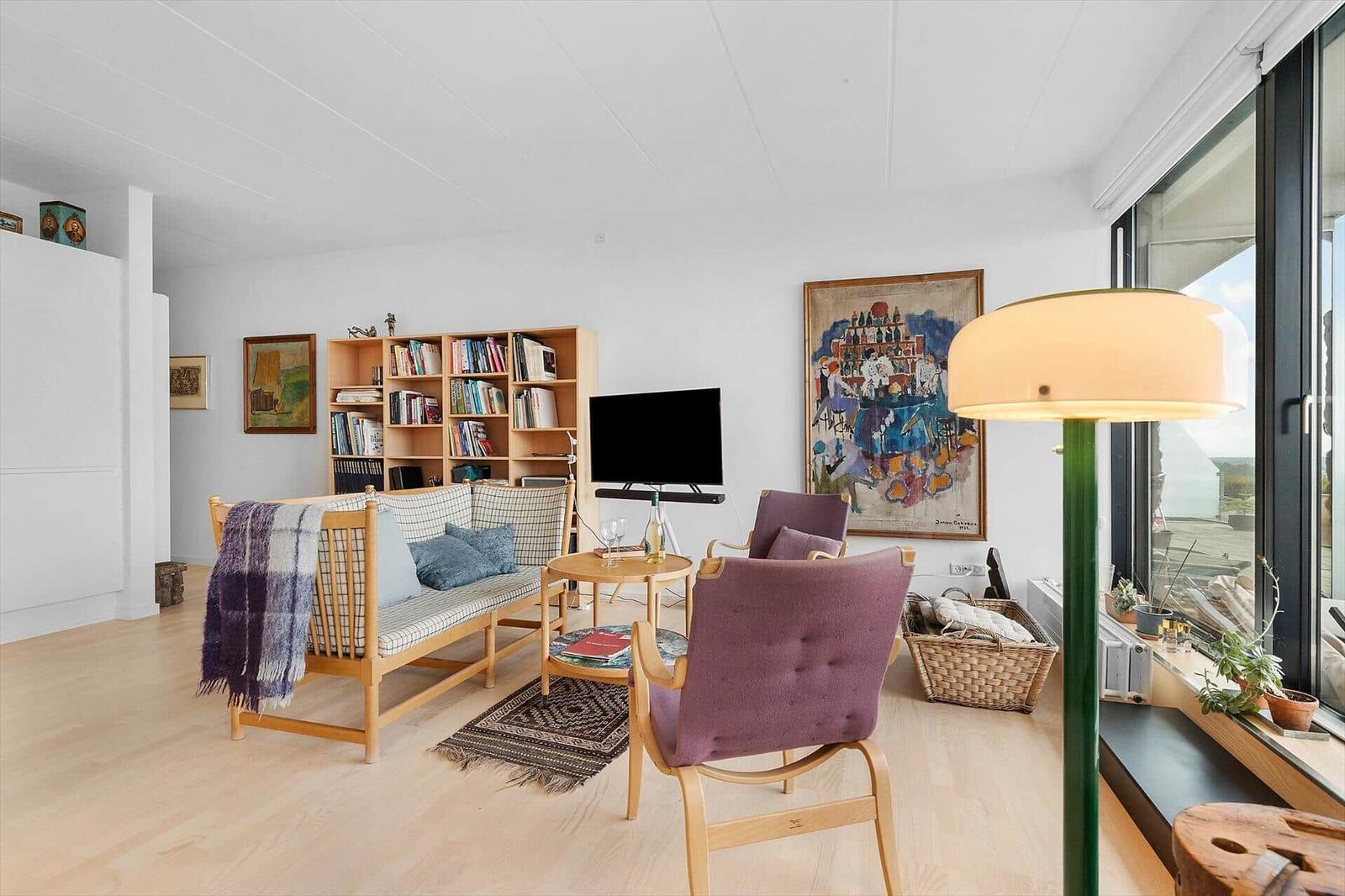 Living room with sofa, chairs, bookshelf, and TV. Large windows and artwork on the walls.
