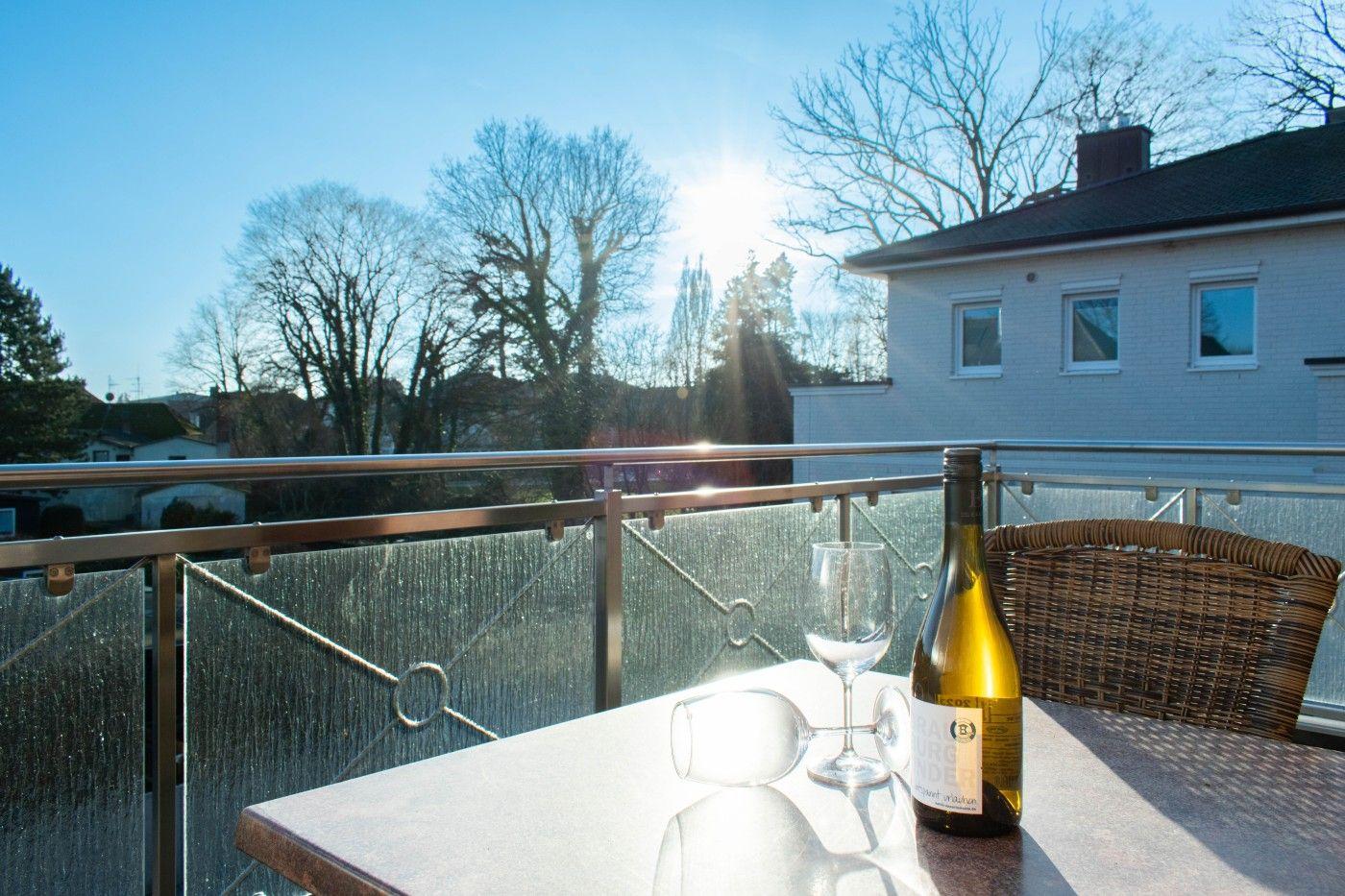 Terrasse mit Tisch, Weinflasche und Gläsern. Hintergrund: Haus und Bäume bei Sonnenschein.