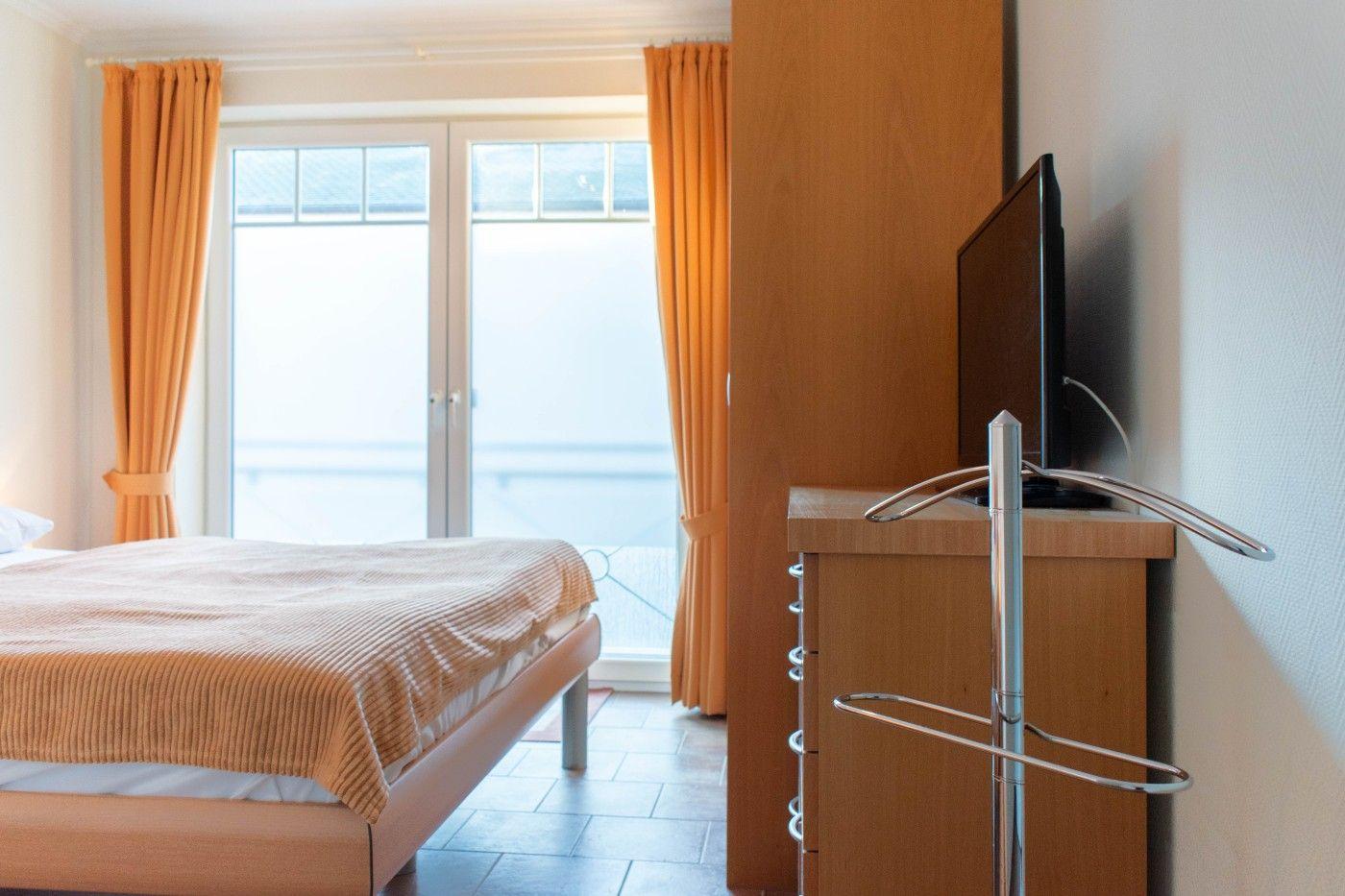 Schlafzimmer mit Bett, TV und Balkontür mit orangefarbenen Vorhängen.