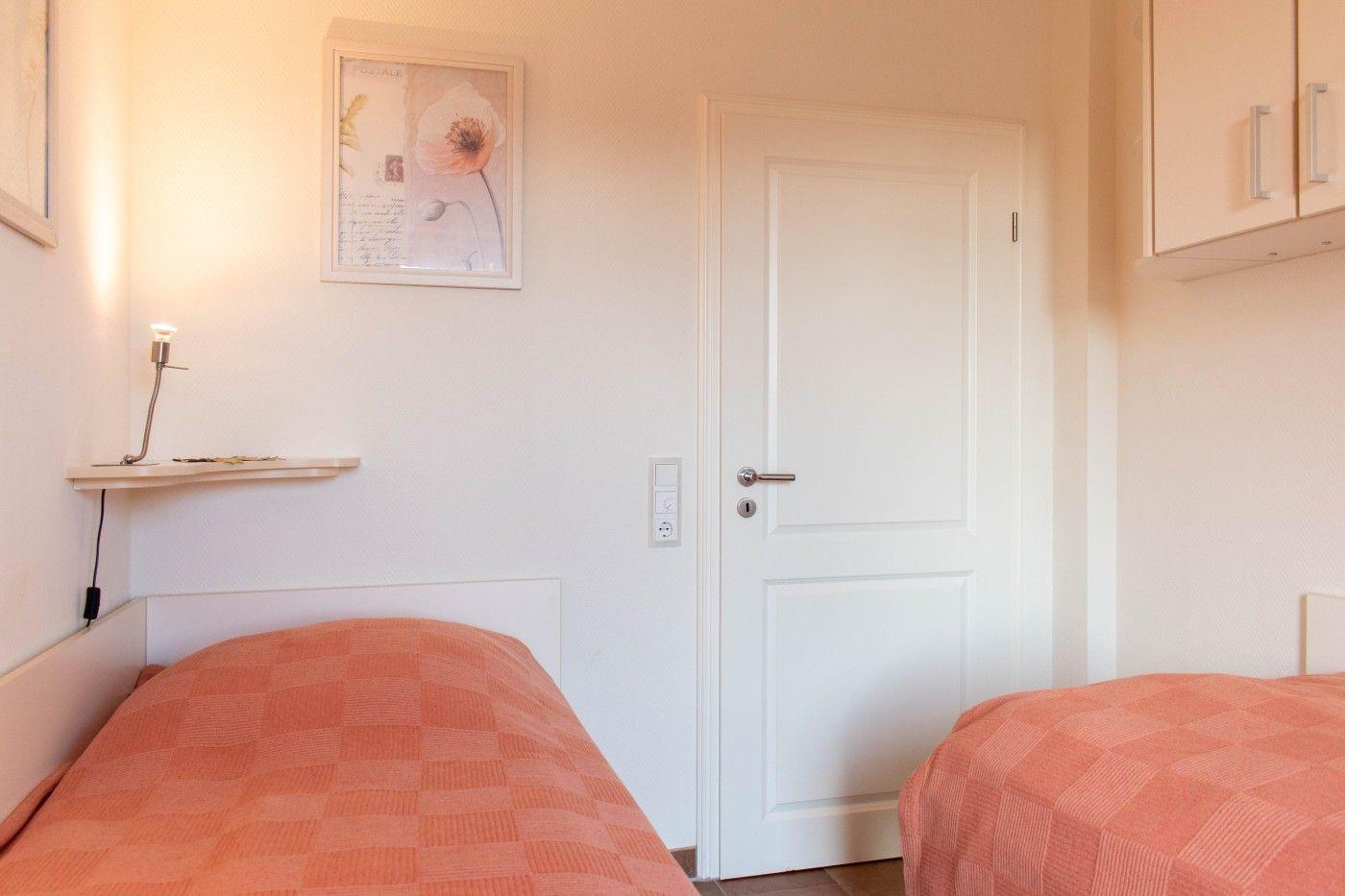 Zwei Betten mit orangefarbenen Bettdecken, weiße Tür und Wandregale mit Bildern.