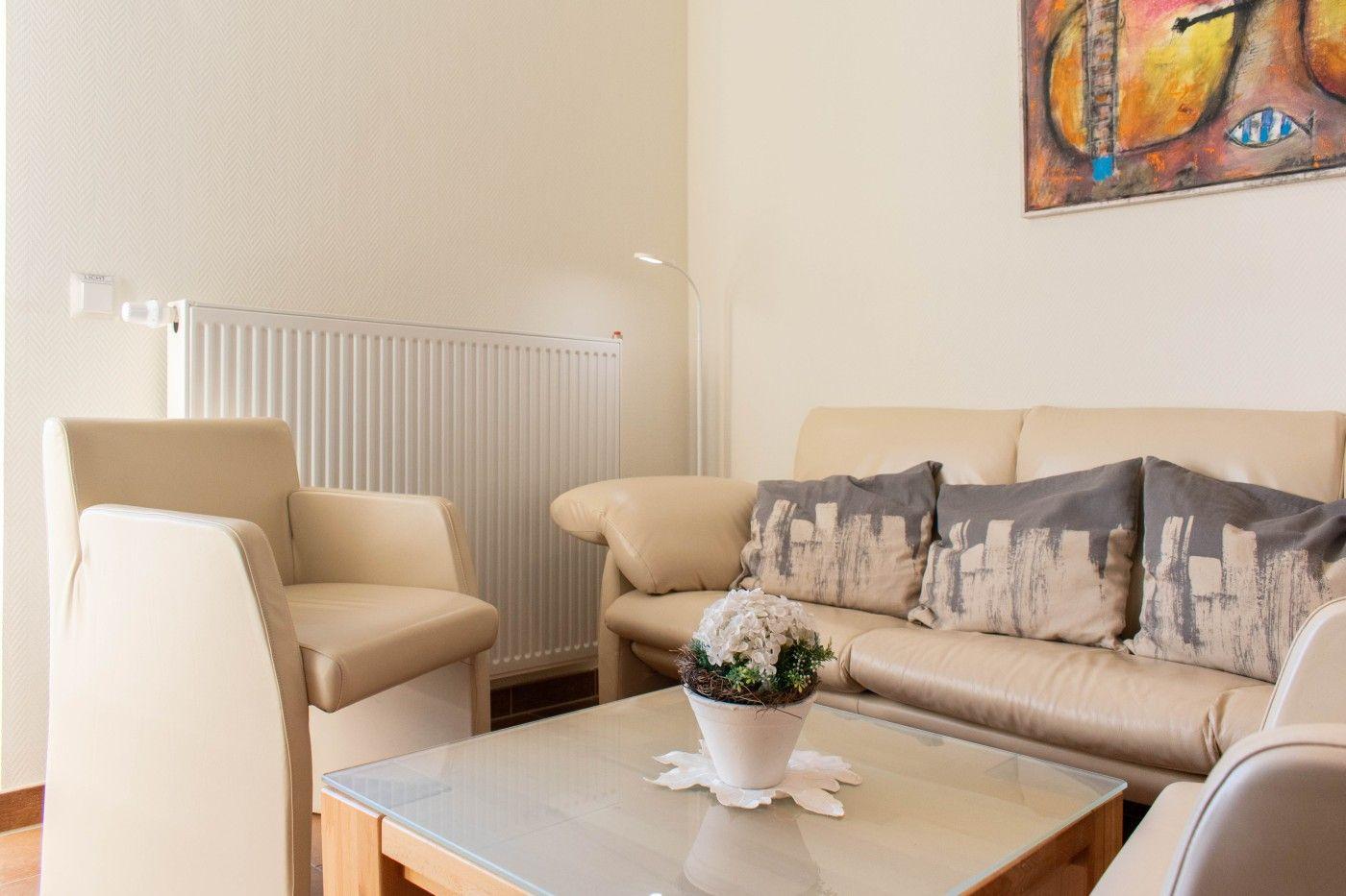 Wohnzimmer mit Sofa, Sessel und Glas-Tisch. Wandbild und Heizkörper sichtbar.