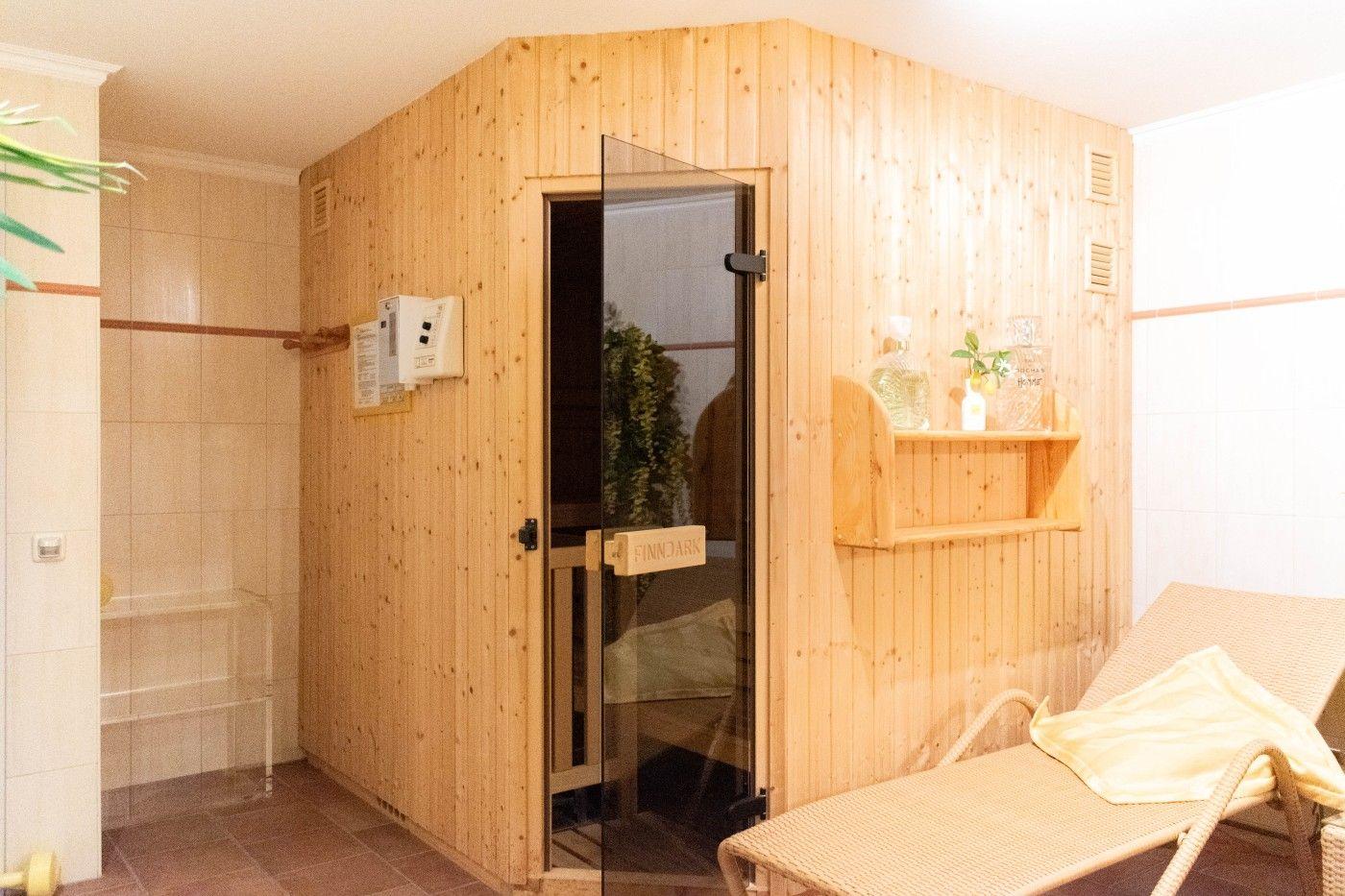 Sauna mit Holzwand, Glasfront und Liegestuhl. Auf Regal stehen Flaschen und Pflanzen.