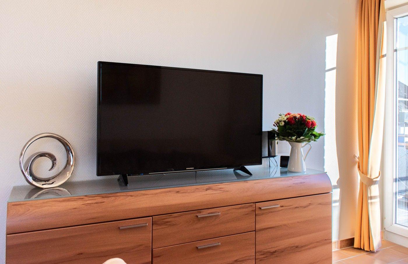 TV auf Holzboard mit Blumen und Dekoration. Fenster mit Vorhängen rechts.