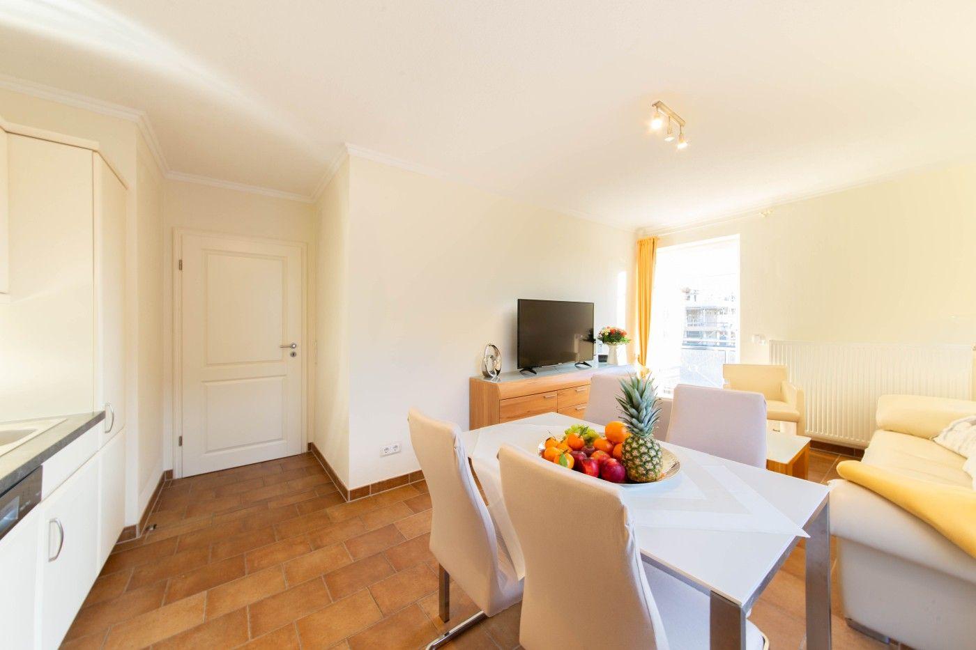 Essbereich mit Tisch, Stühlen und Obst. Wohnzimmer mit Sofa und TV. Küche mit Arbeitsplatte.